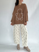 Person wearing a brown sweater with a gingerbread man design and white pants with brown polka dots on a plain background