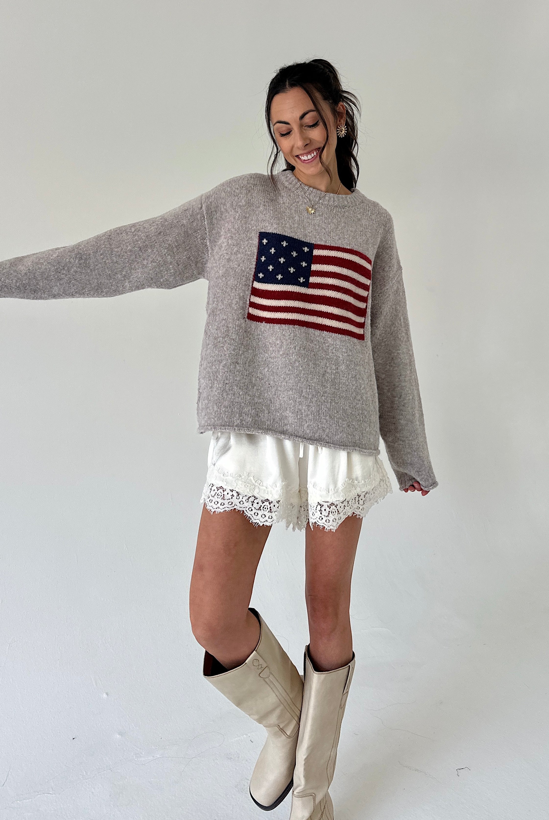 A woman modeling a light gray knit sweater with an American flag graphic on the chest, paired with white lace-trim shorts and beige cowboy boots, against a plain light gray studio background.