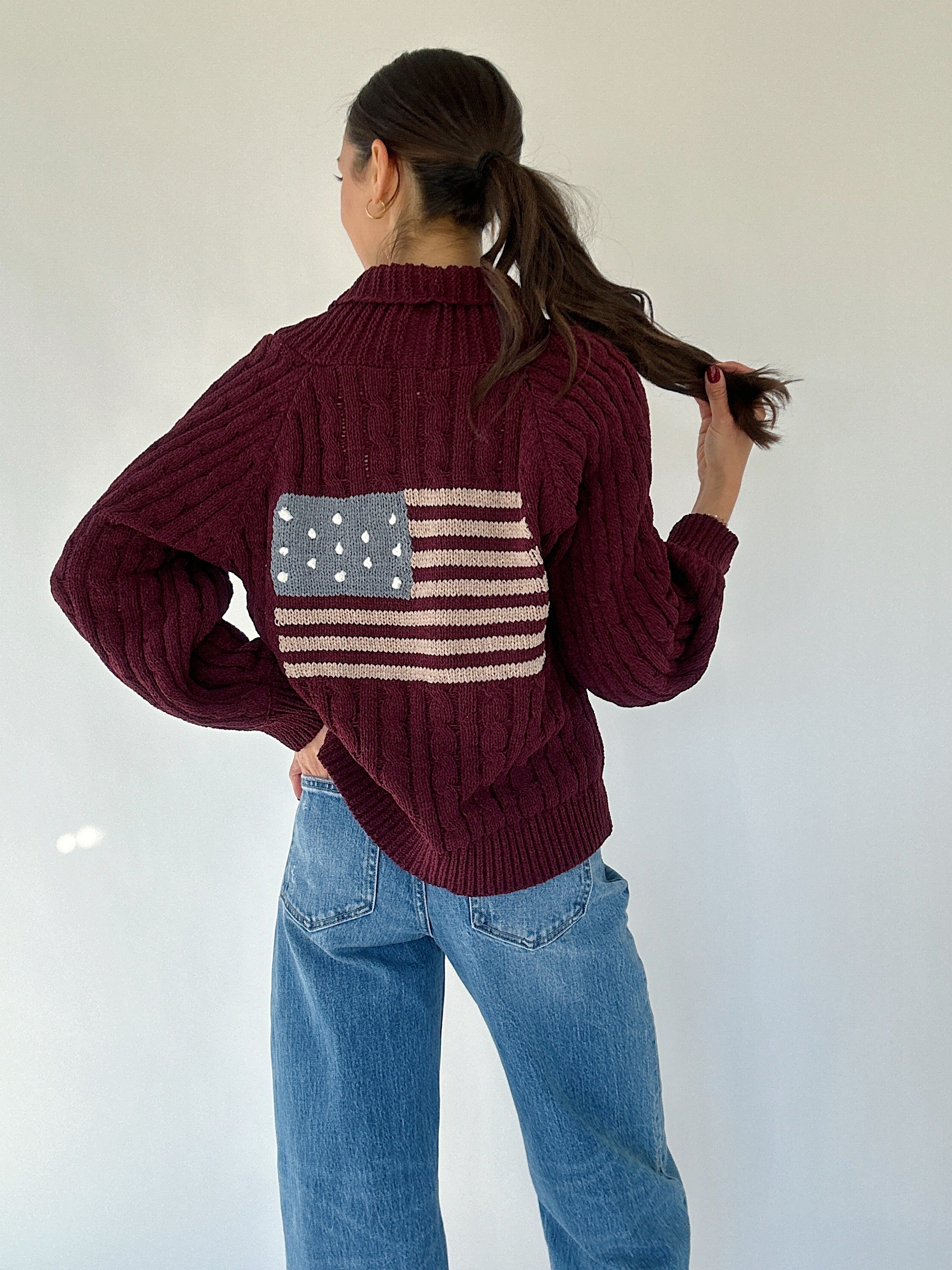 Person wearing a maroon sweater with an American flag design on the back on a white background