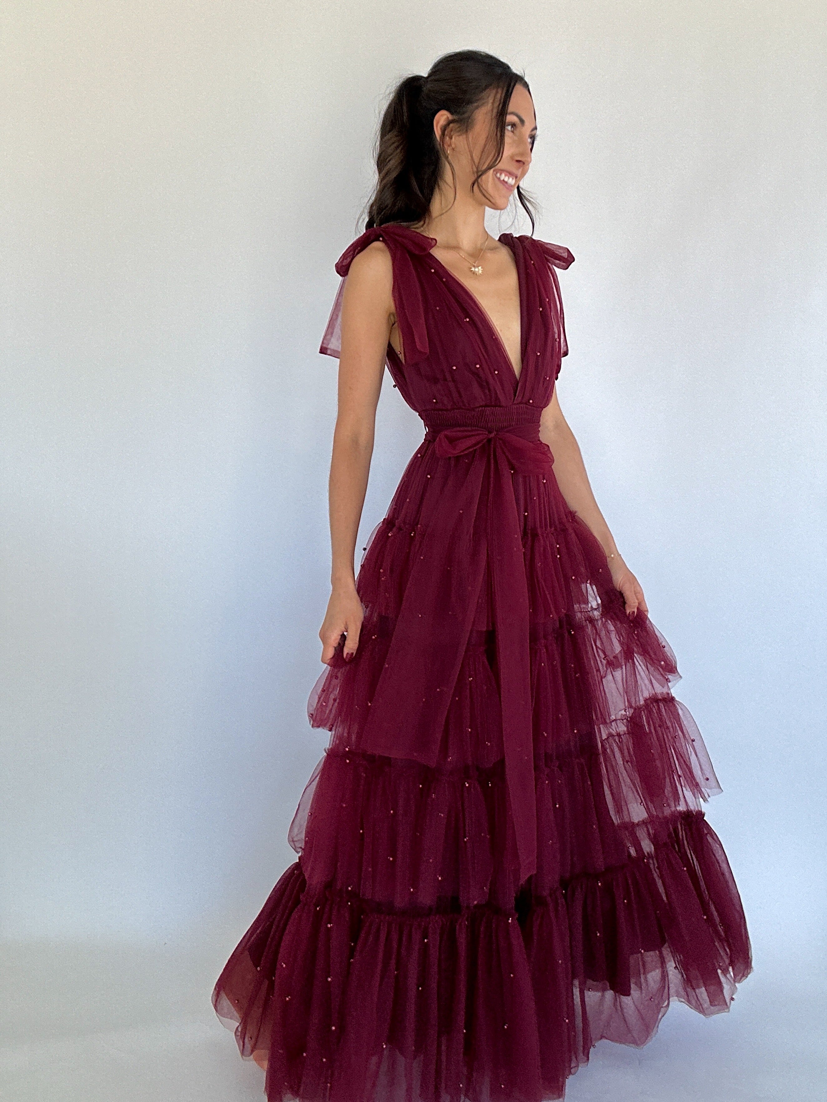 Woman wearing a burgundy evening gown with a white background holding tiered skirt