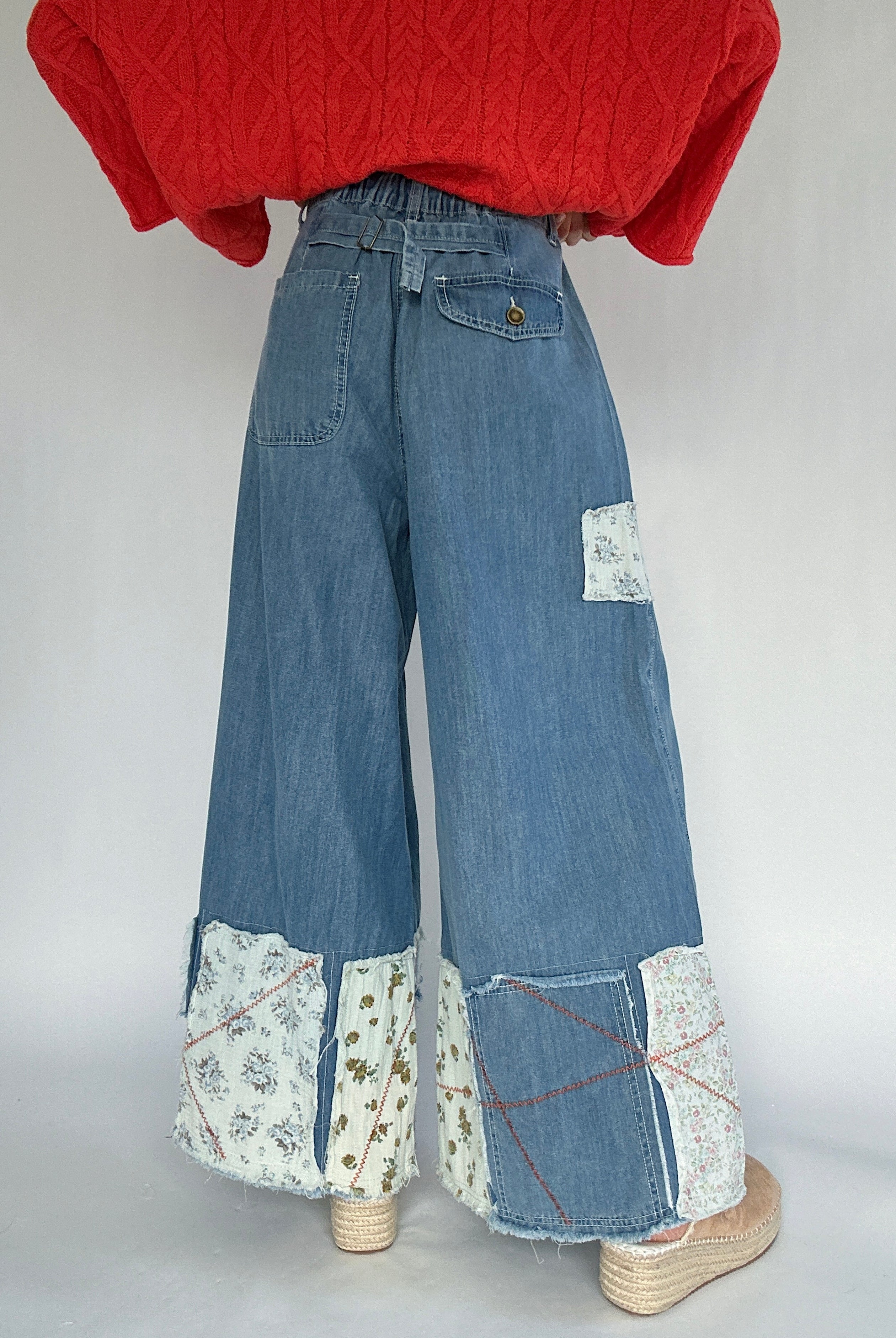 Person wearing a coral sweater and blue jeans with patchwork details a back buckle and back pockets on a plain background