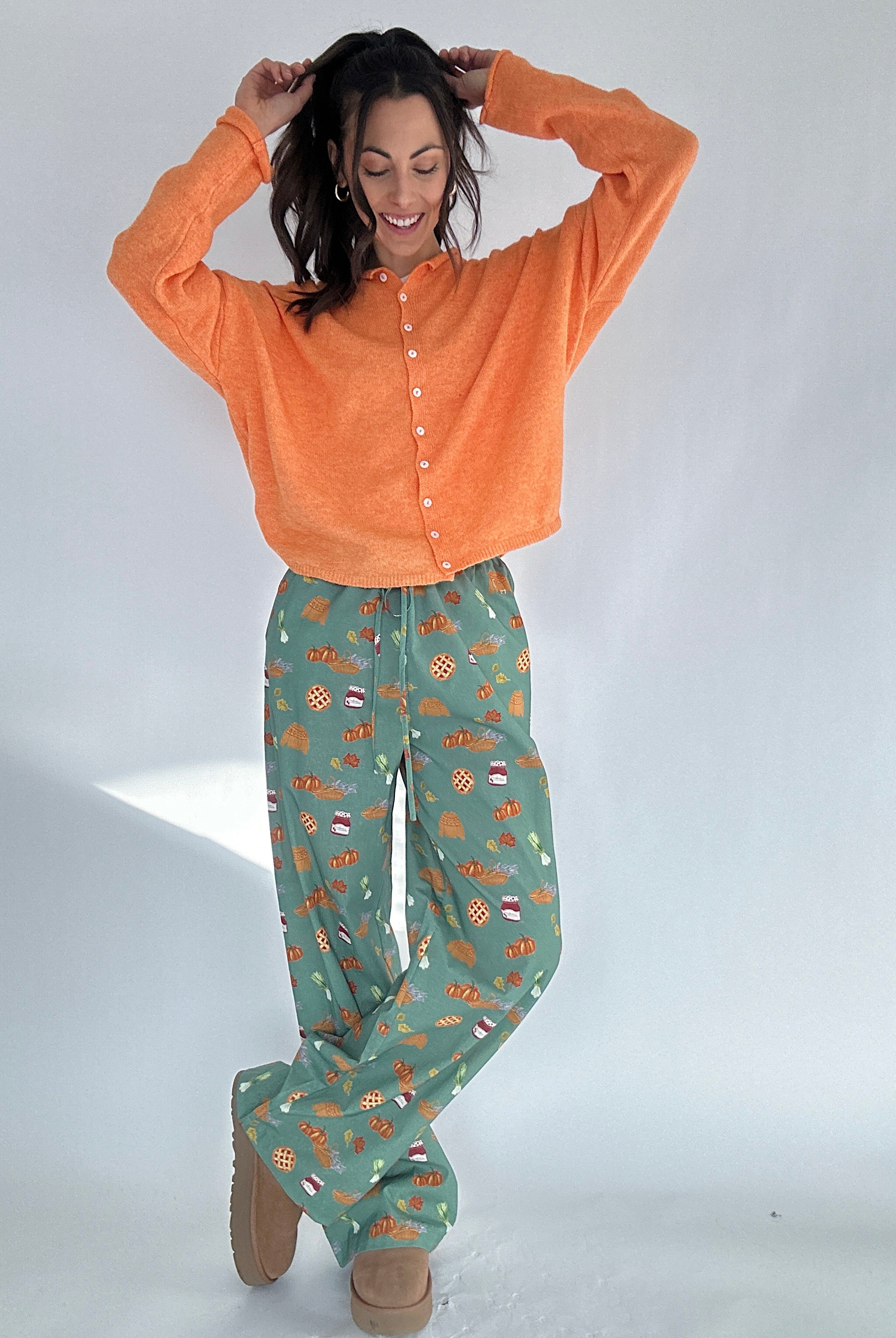 Woman wearing an orange cardigan sweater and green fall patterned pants on a white background