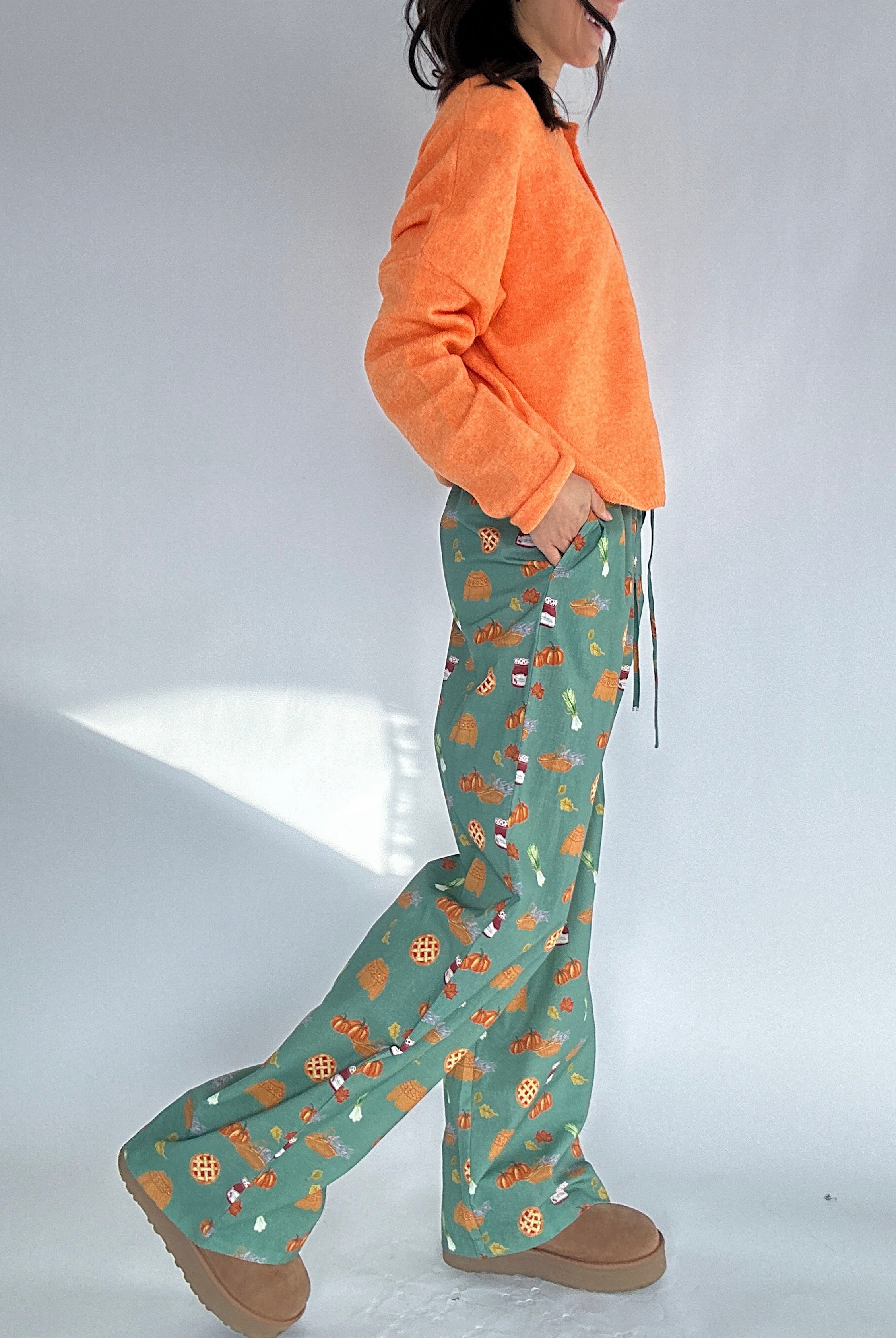 Person wearing an orange cardigan top and green fall patterned pants on a white background showing side view with pockets