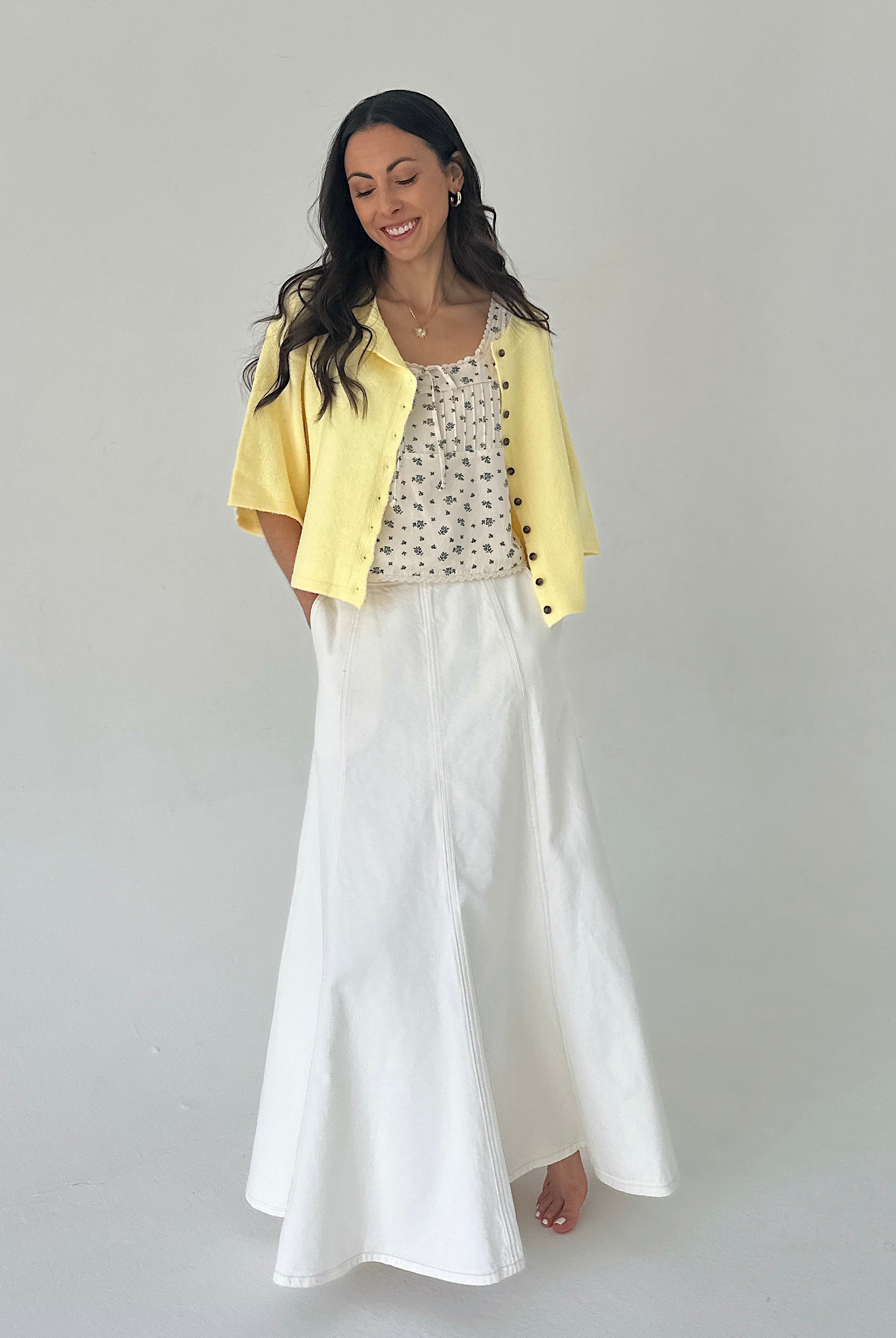 Woman wearing a yellow cardigan, patterned top, and white skirt on a plain background
