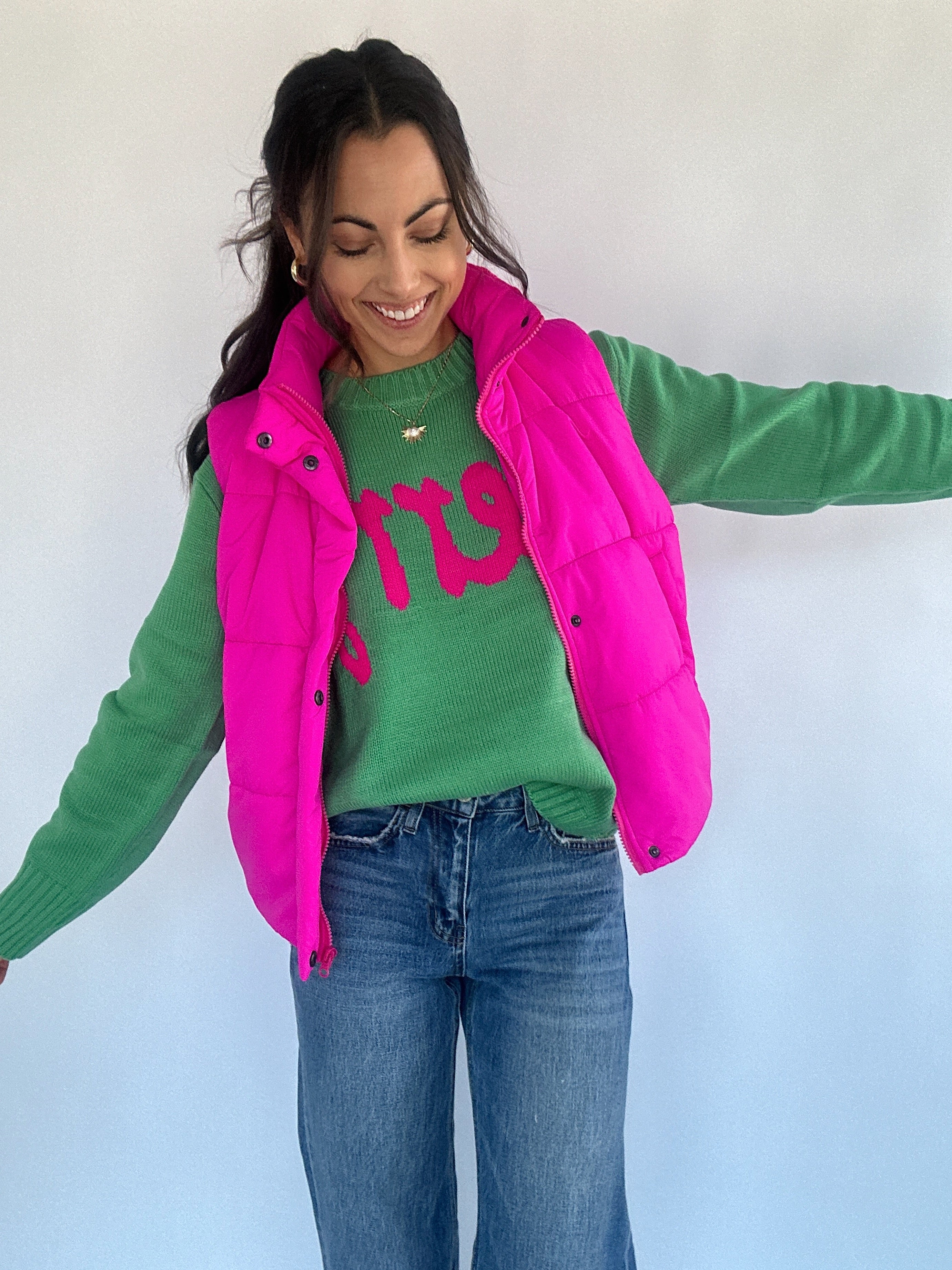 Woman wearing green sweater with text under a hot pink puffer vest and jeans on a plain background
