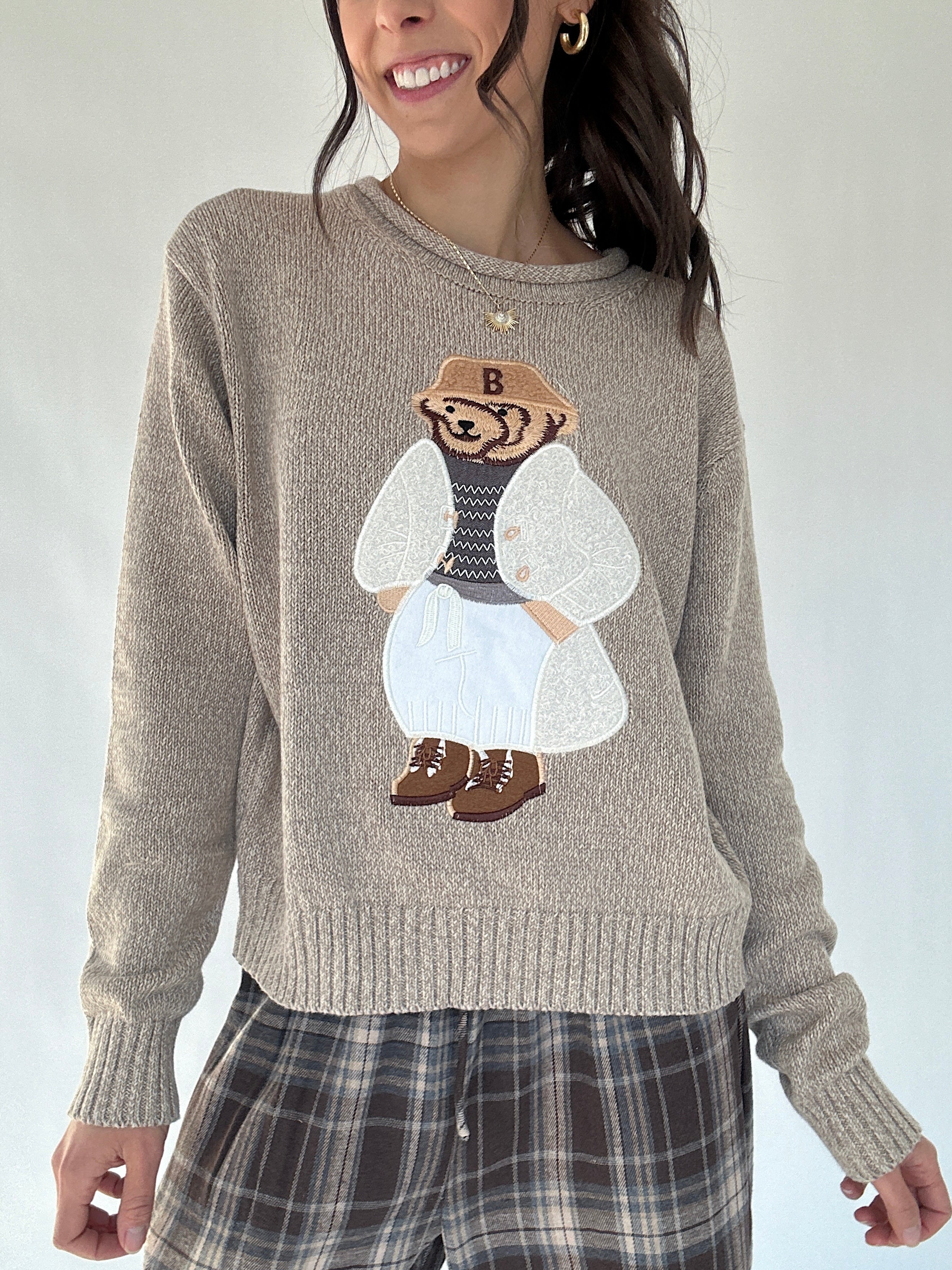 Person wearing a taupe sweater with a dressed up teddy bear embroidered character design on a plain background