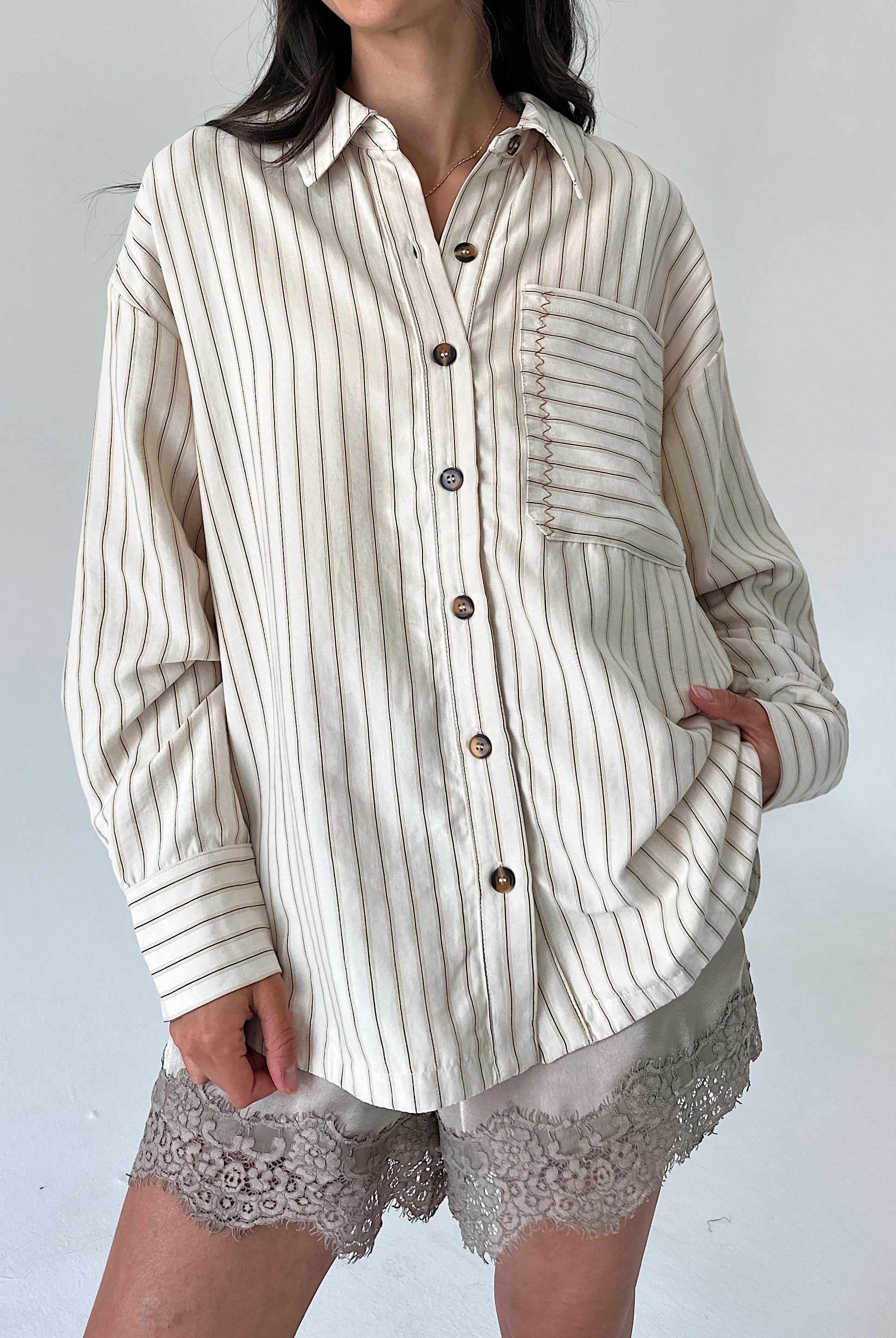 A woman wearing an oversized cream-colored button-up shirt with thin vertical brown stripes, paired with lace-trimmed shorts, against a plain light gray background.