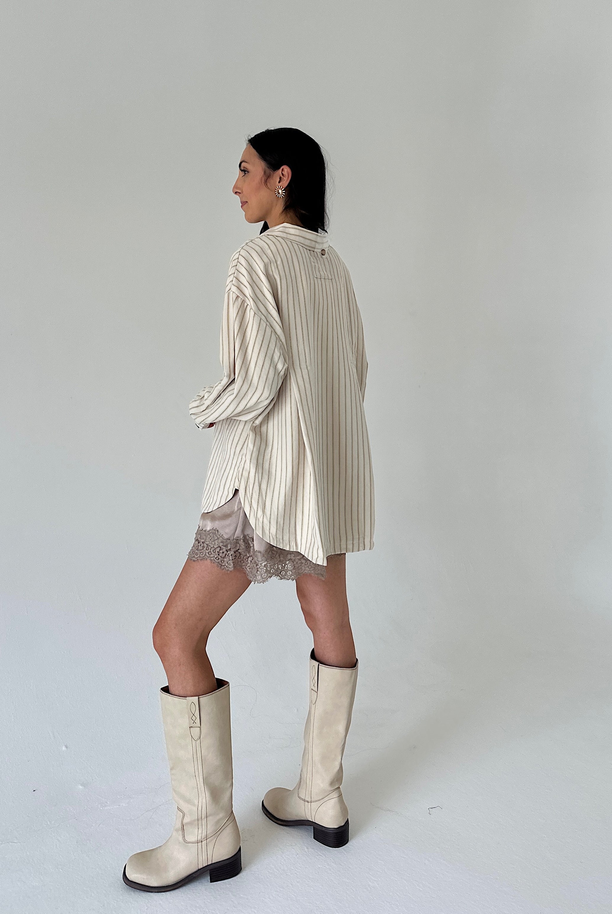 A woman is shown from the back wearing a cream and beige vertically striped oversized shirt dress, a sheer lace mini skirt, and off-white knee-high boots against a plain light gray studio background.
