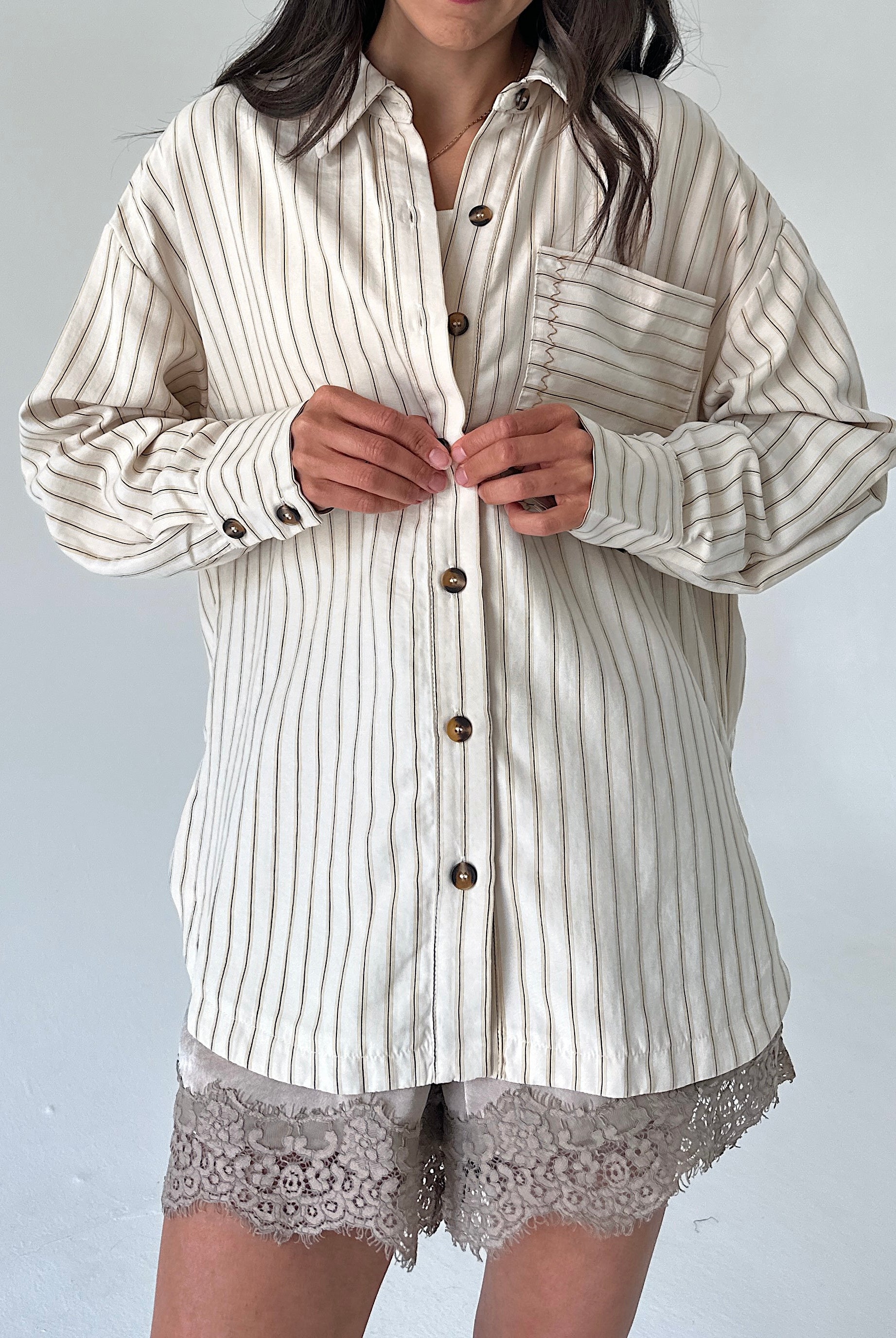 A woman wearing an oversized cream-colored button-up shirt with thin vertical brown stripes, paired with lace-trimmed shorts, against a plain light gray background.