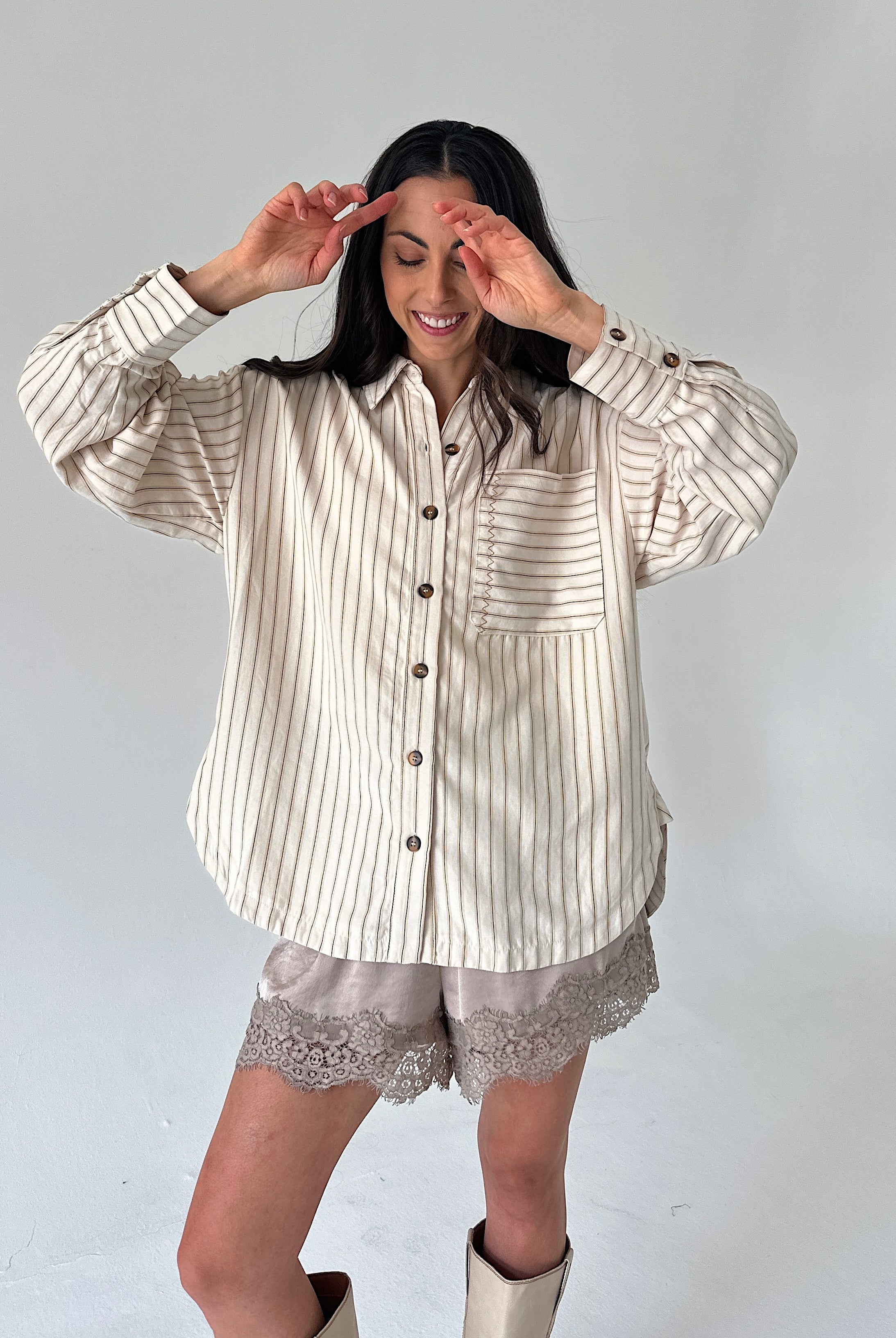 A woman modeling a cream and brown vertically striped button-up shirt with long sleeves, paired with matching lace-trim shorts and beige boots, against a plain light gray studio background.