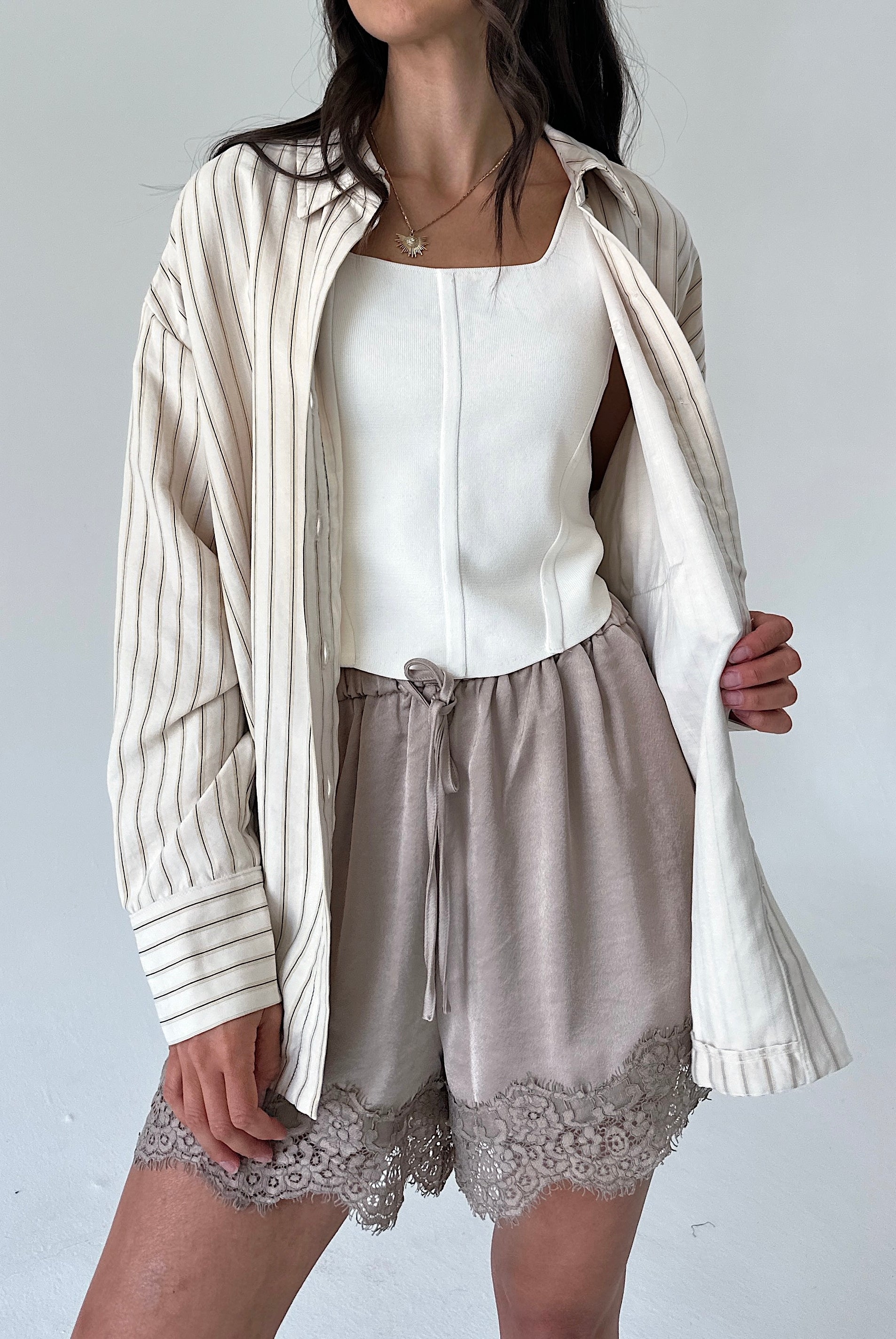 A model wearing a cream and beige vertically striped button-up shirt draped open over a white camisole, paired with taupe drawstring shorts featuring a lace hem.