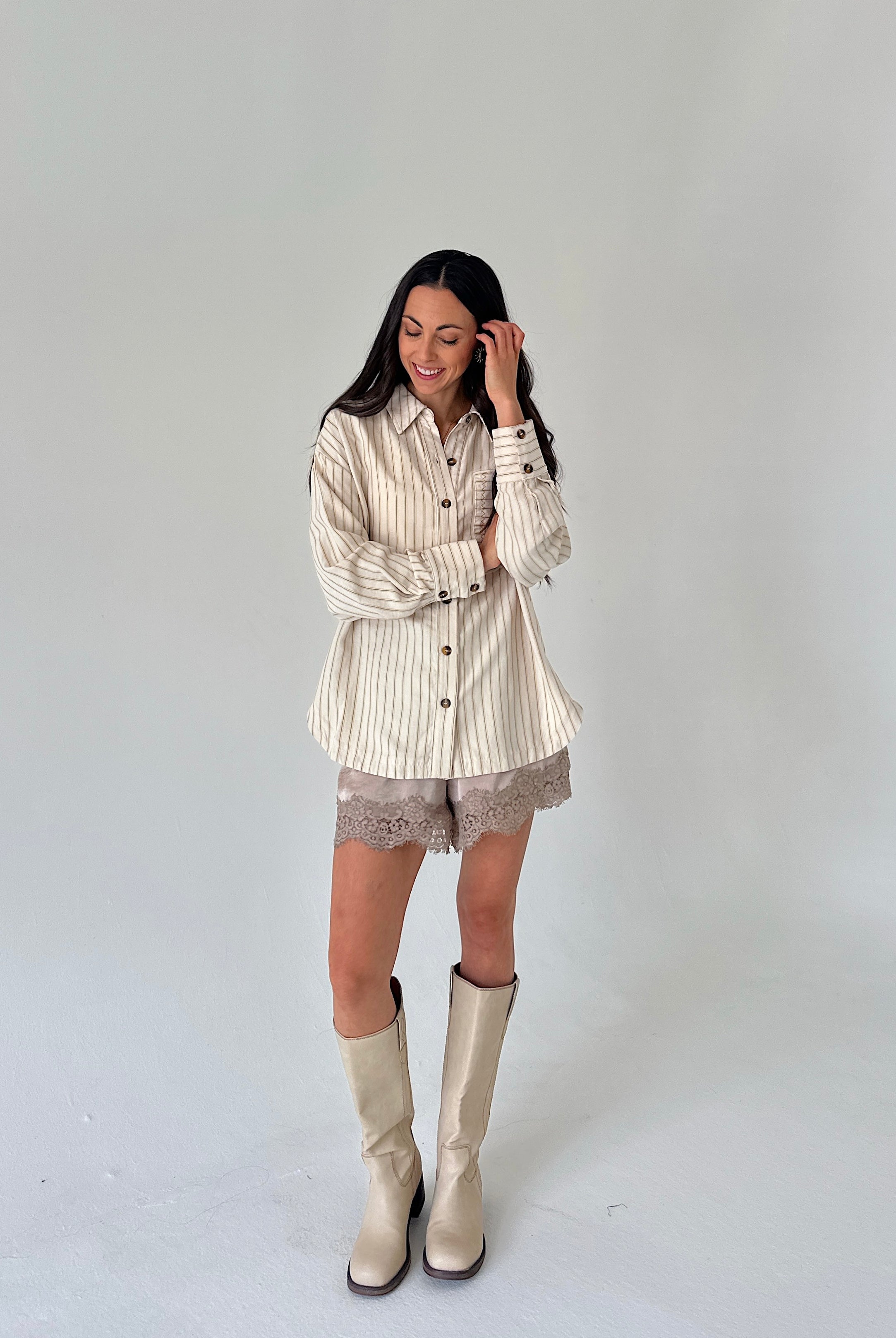 A woman modeling a cream and beige vertically striped button-down shirt with lace shorts and beige knee-high boots, standing against a plain light gray studio background.