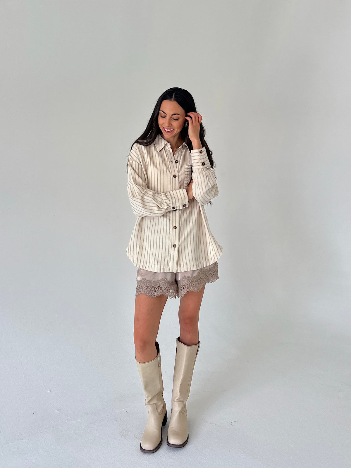 A woman modeling a cream and beige vertically striped button-down shirt with lace shorts and beige knee-high boots, standing against a plain light gray studio background.