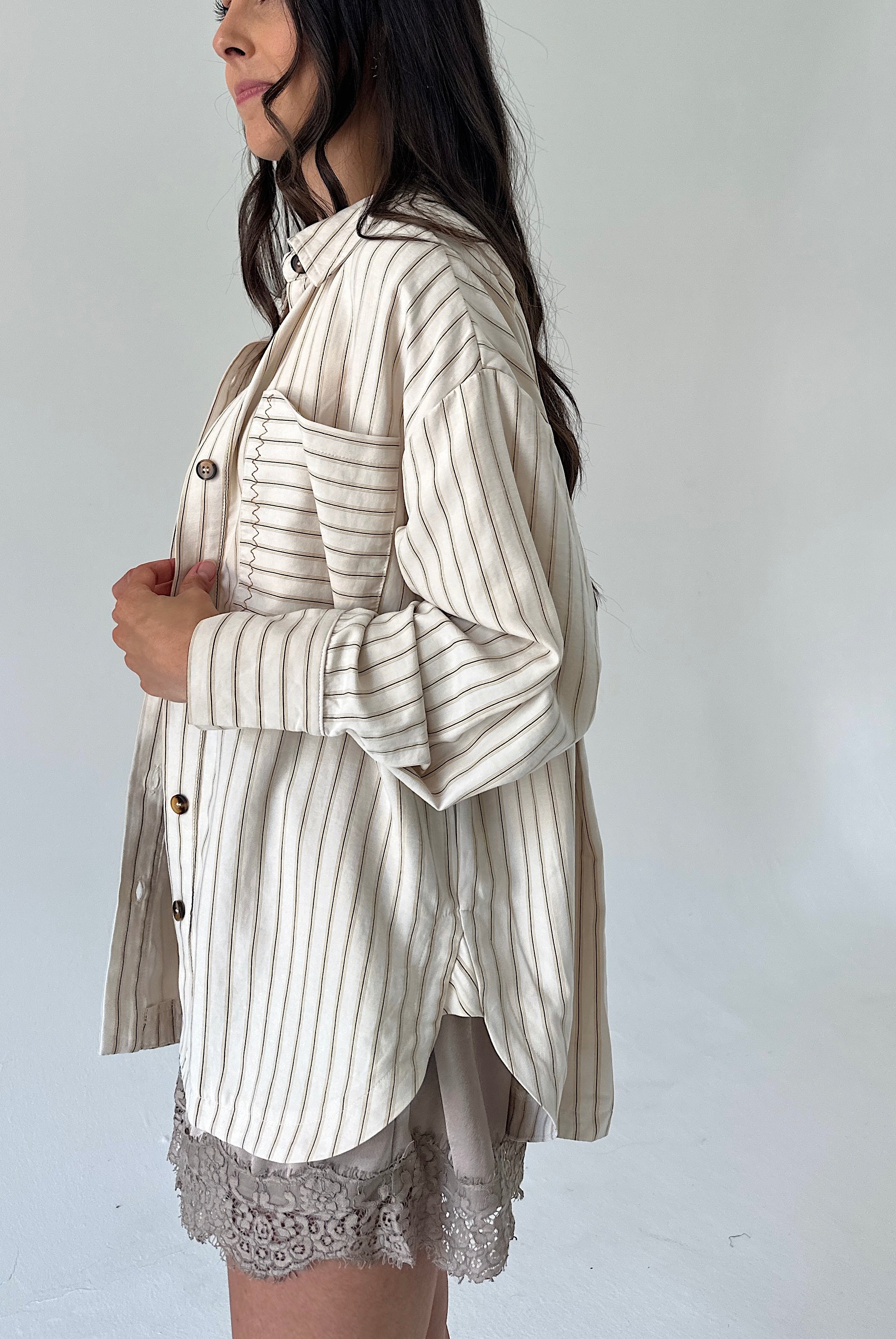 A woman wearing a cream-colored button-up shirt with thin vertical brown stripes, styled over a light gray lace-trimmed skirt, shown from a side profile against a plain light gray background.