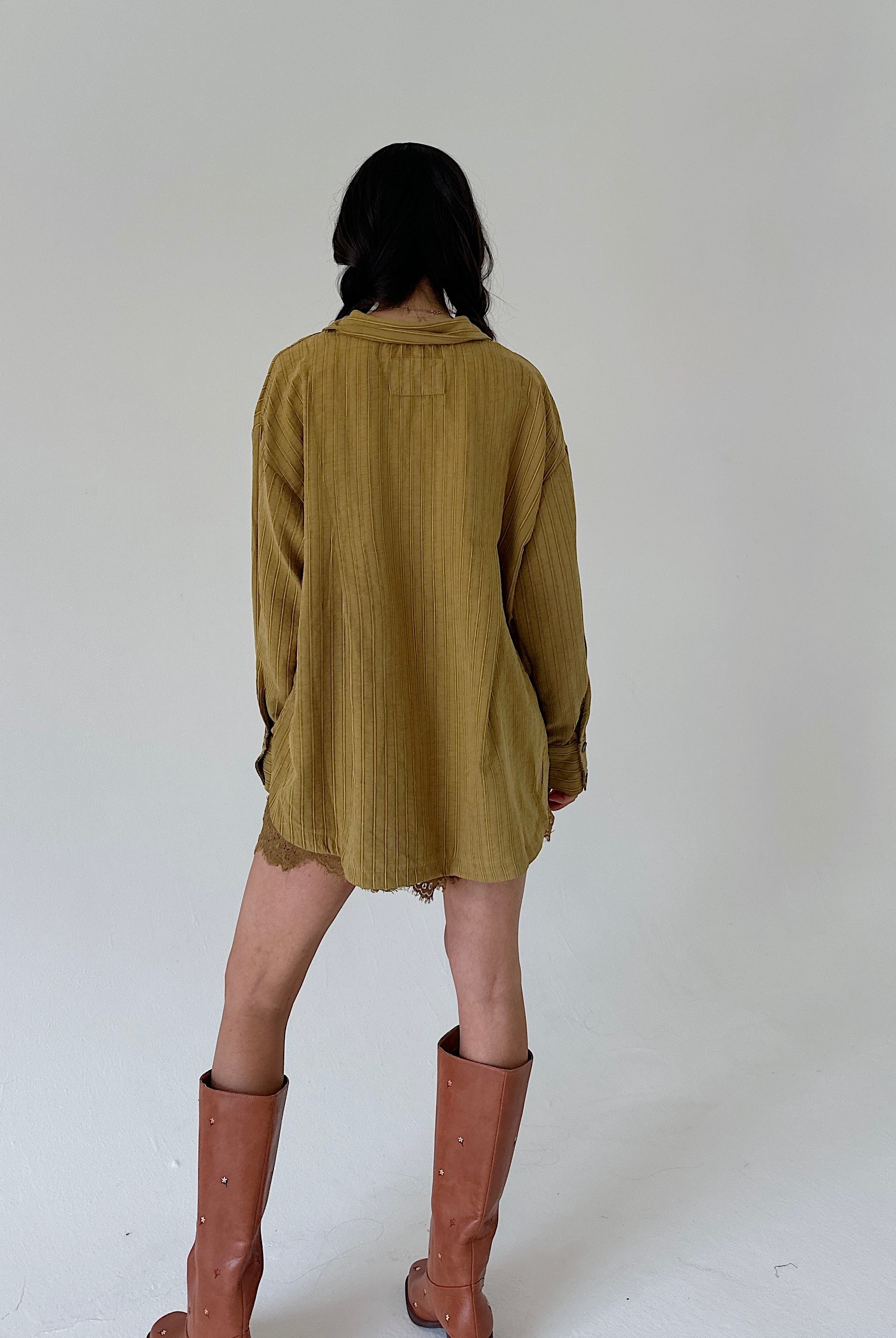 A model viewed from behind wearing a loose, olive-yellow shirt and brown leather knee-high boots against a plain white studio background.
