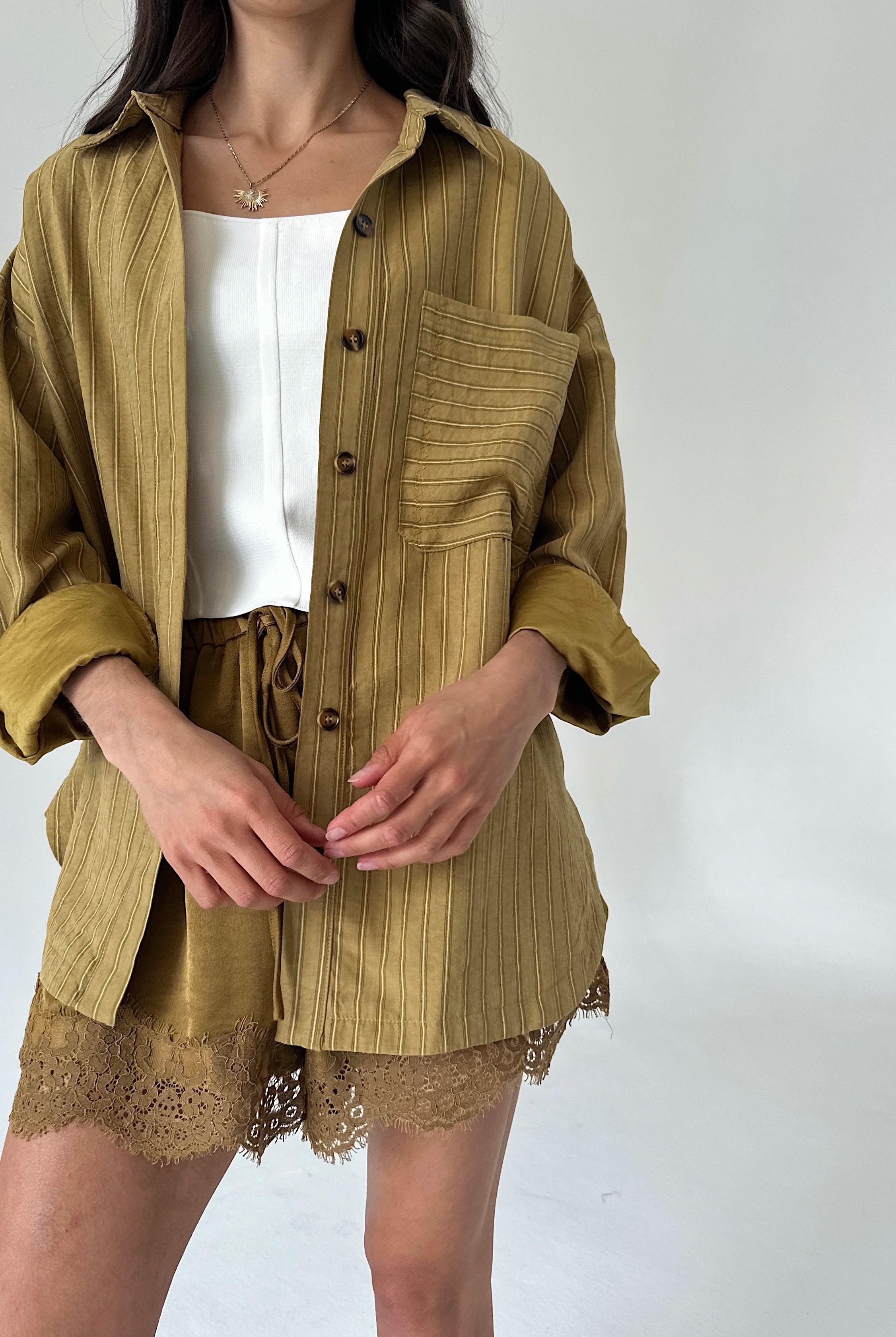 A model wearing a matching olive mustard button up shirt and shorts set with a white top underneath, photographed against a plain light gray background.