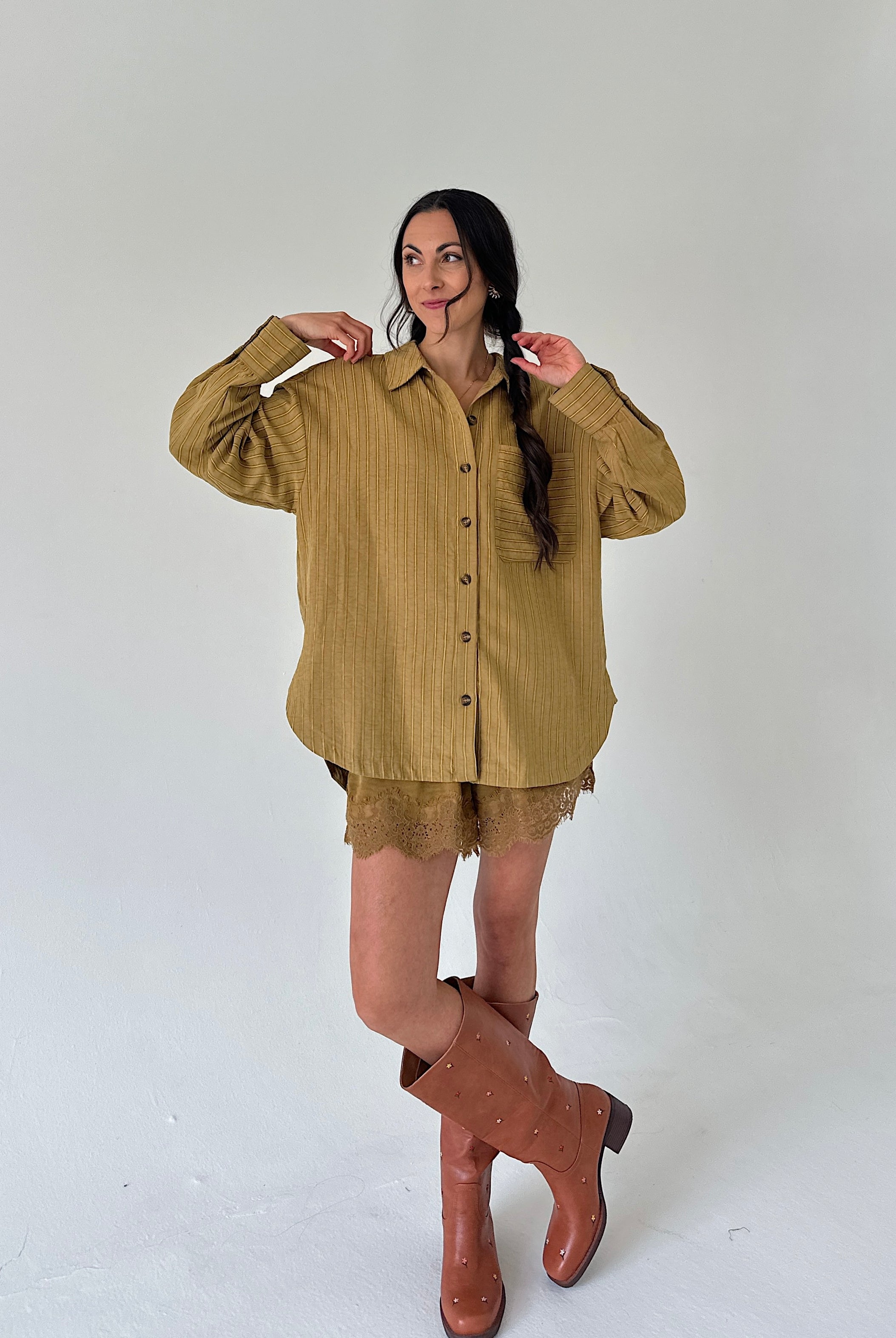 A woman modeling an oversized olive-brown linen button-up shirt with matching lace shorts and brown leather knee-high boots, standing against a plain light gray studio background.