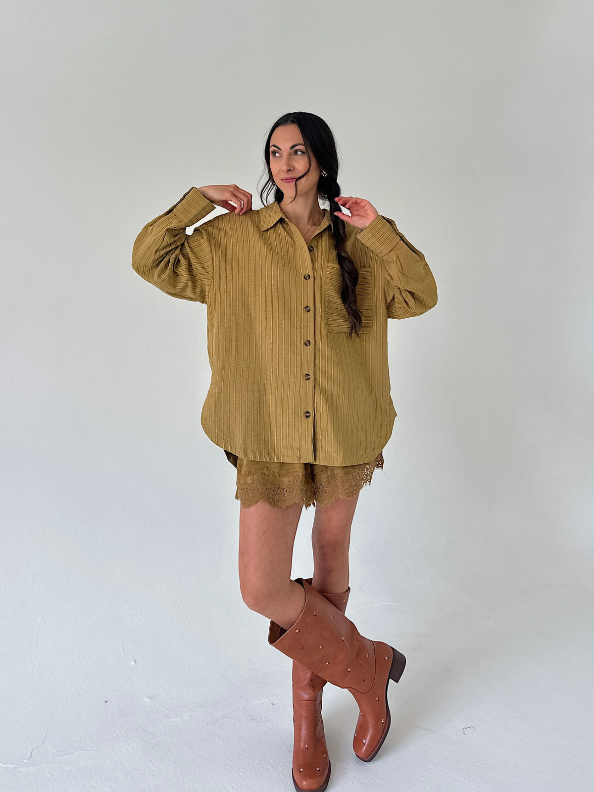 A woman modeling an oversized olive-brown linen button-up shirt with matching lace shorts and brown leather knee-high boots, standing against a plain light gray studio background.