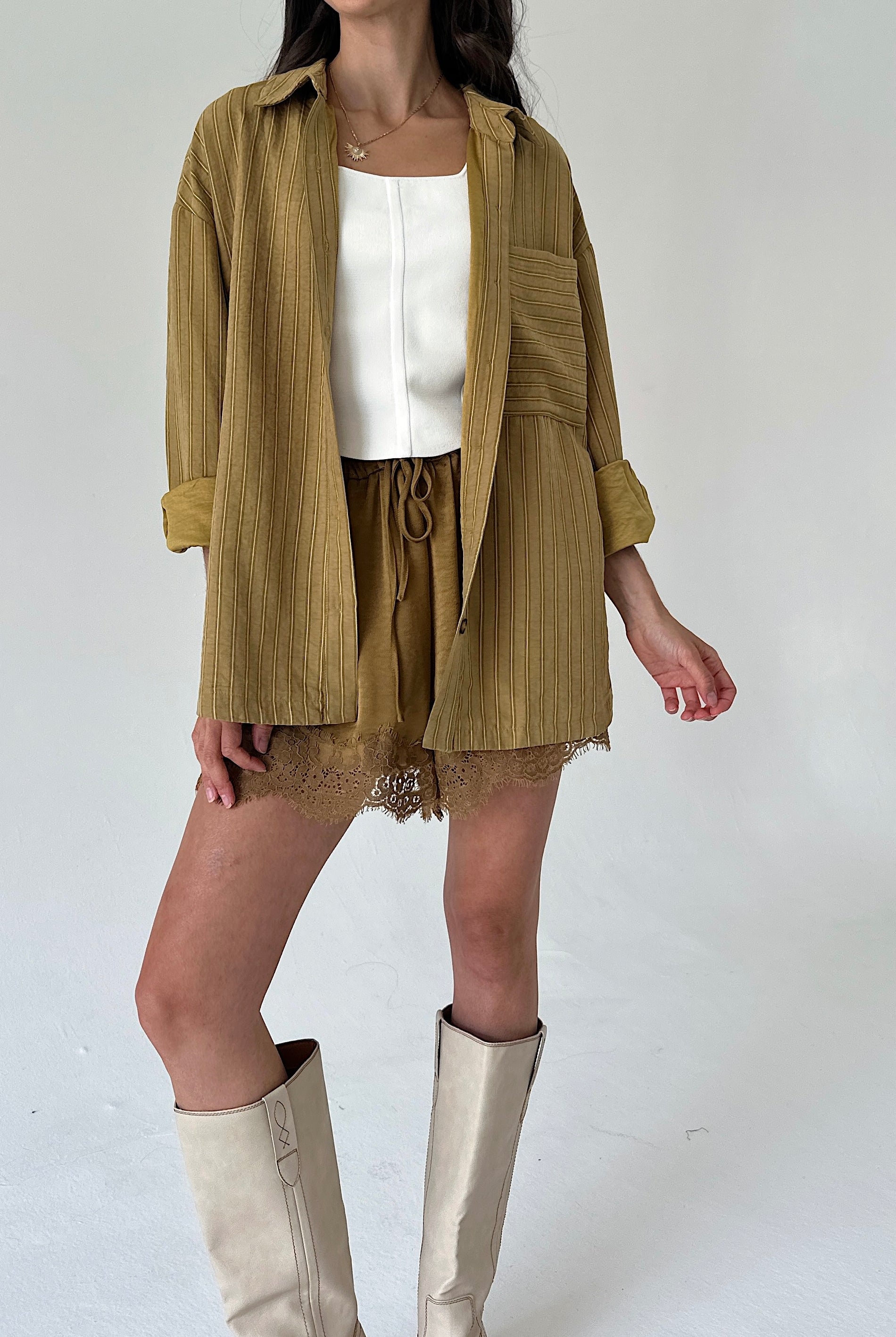 A model wearing a mustard yellow shirt jacket over a white top, paired with matching satin shorts and beige knee-high boots, against a plain light gray background.
