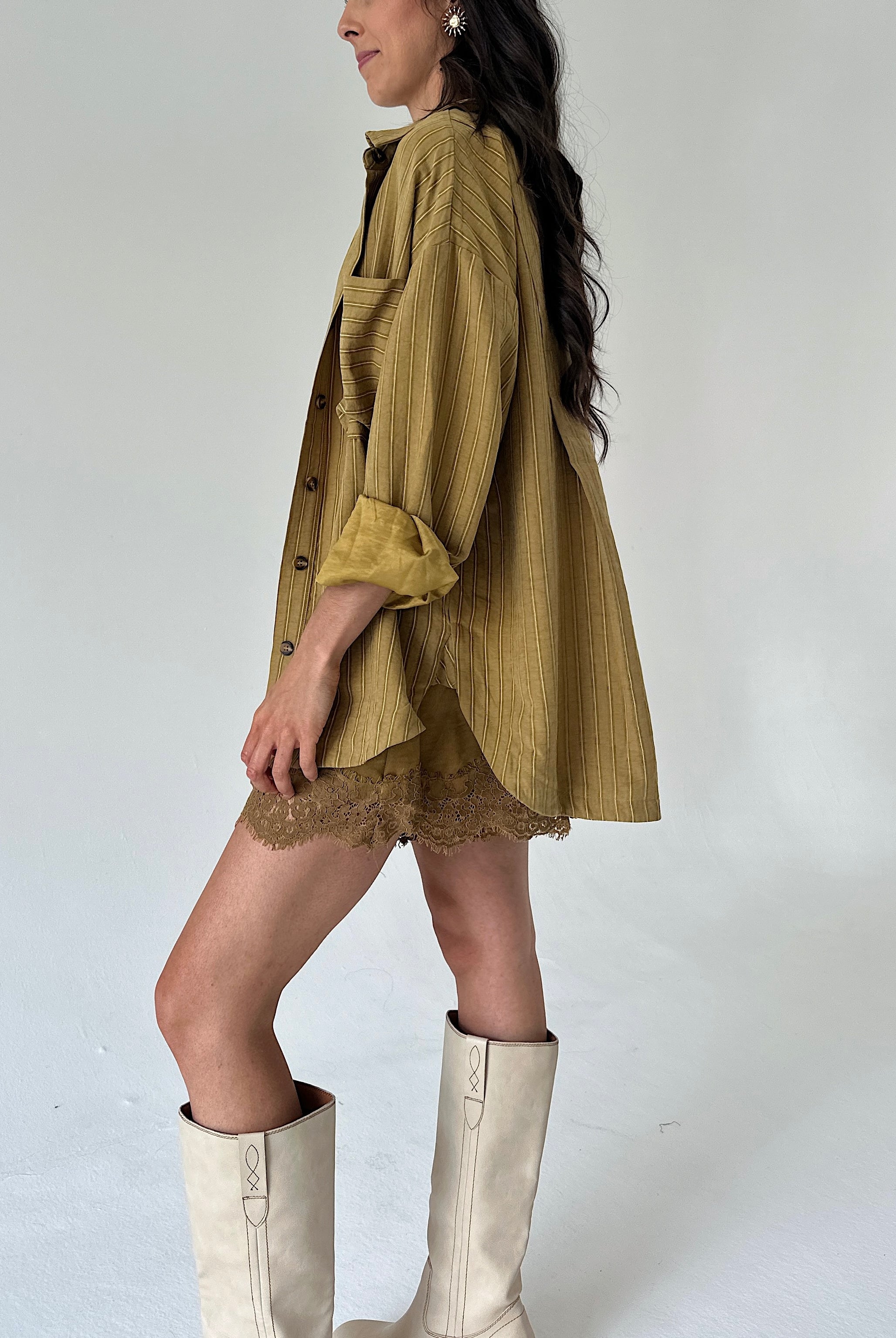 A woman wearing an oversized mustard yellow shirt with a lace hem shorts, styled with cream-colored knee-high boots against a plain light gray studio background.