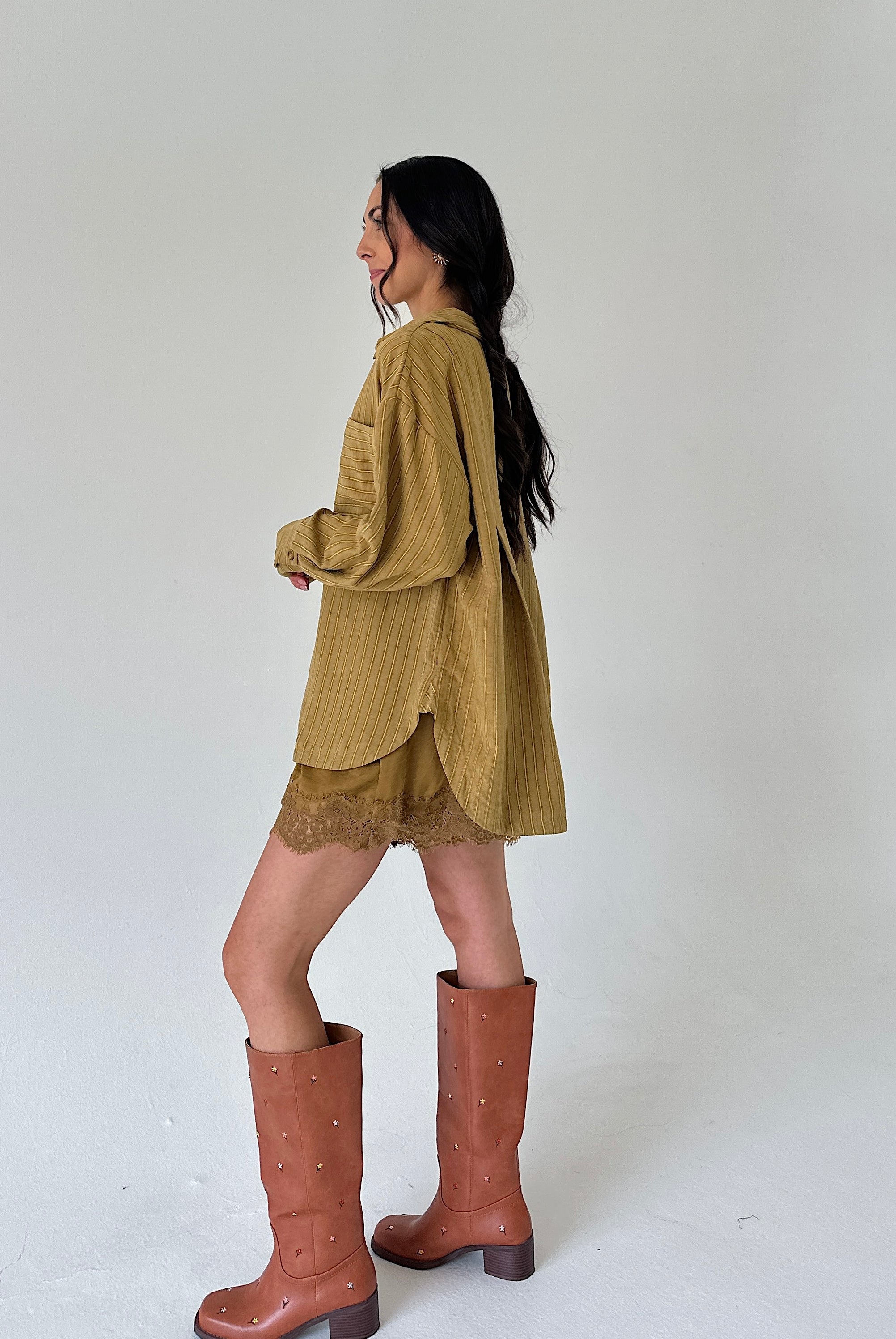 A woman is shown from the side wearing a mustard yellow shirt, matching lace hem shorts, and brown studded knee-high boots against a plain white studio background.