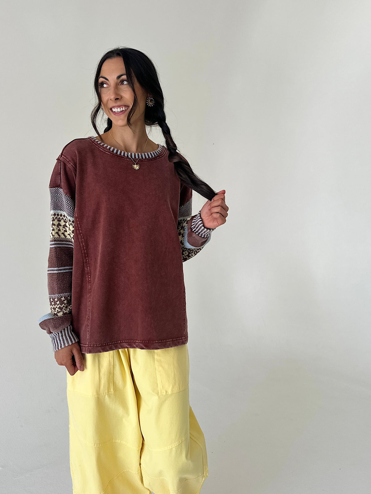 A woman modeling a rust-brown long-sleeve top with patterned knit sleeves and yellow cargo pants, standing against a plain off-white wall.