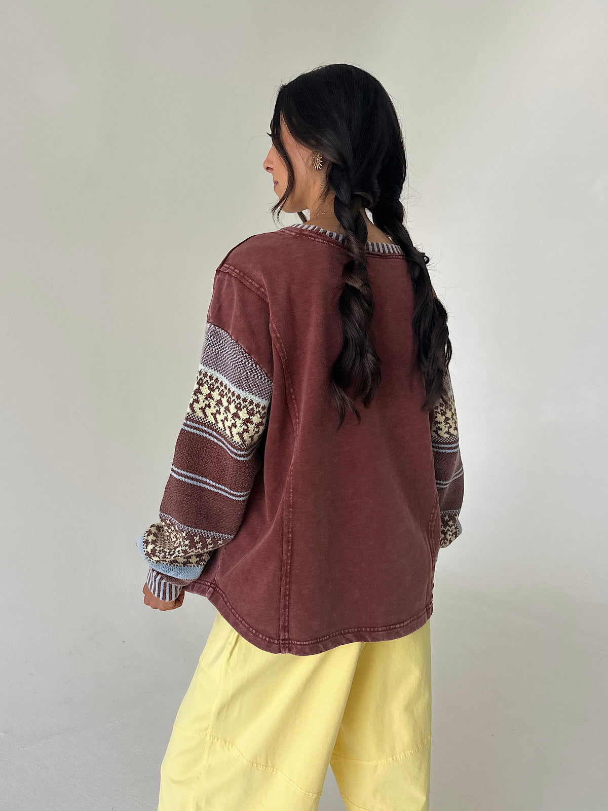 A woman with long dark hair in a braid is shown from the back, wearing a washed reddish-brown cropped sweatshirt with patterned knit sleeves and yellow pants, standing against a plain light gray wall.
