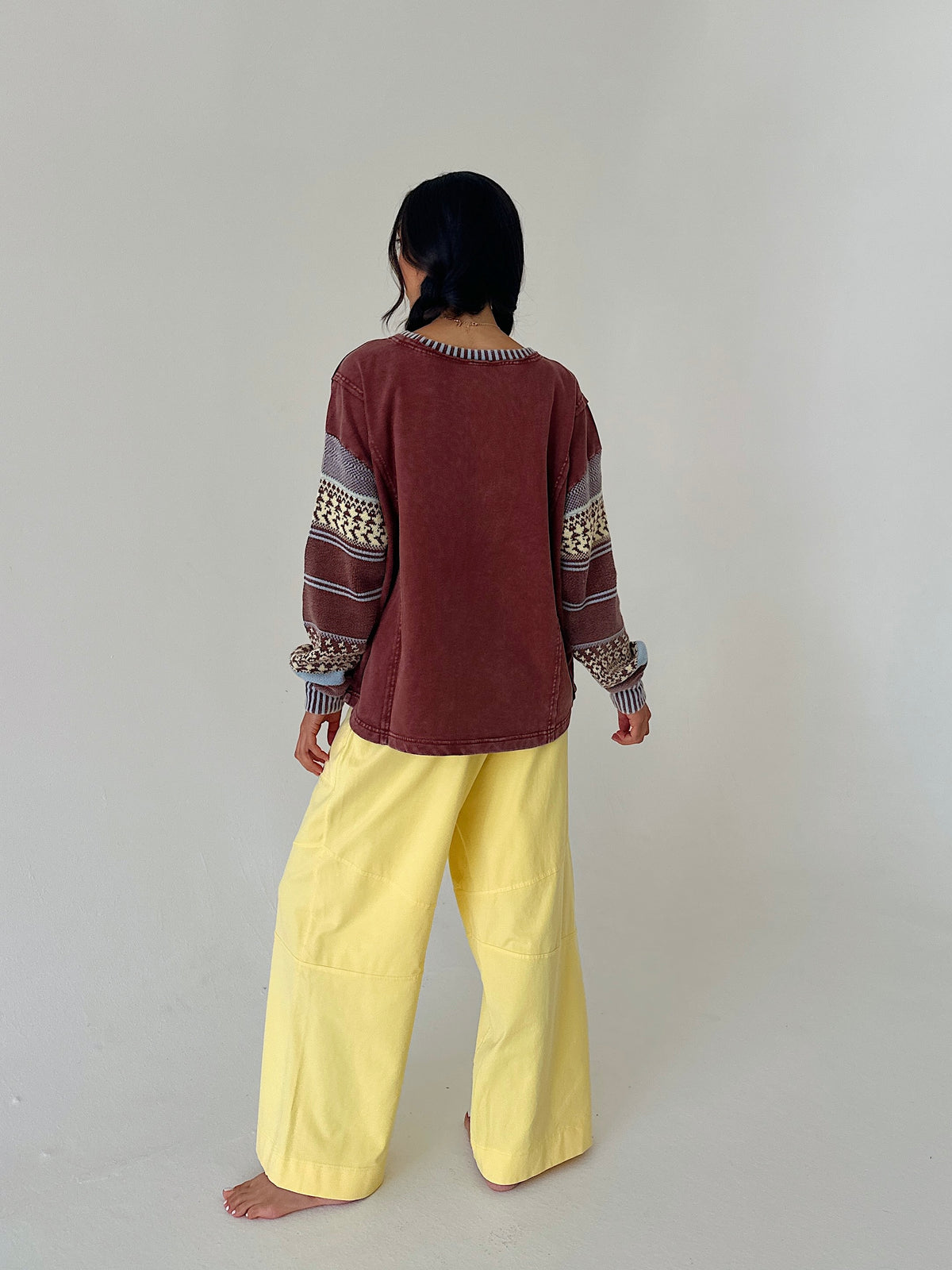 A person standing barefoot against a plain light gray studio background, shown from the back. They are wearing a loose long-sleeve top in dark reddish brown with patterned knit cuffs and shoulders, paired with wide-leg light yellow pants.
