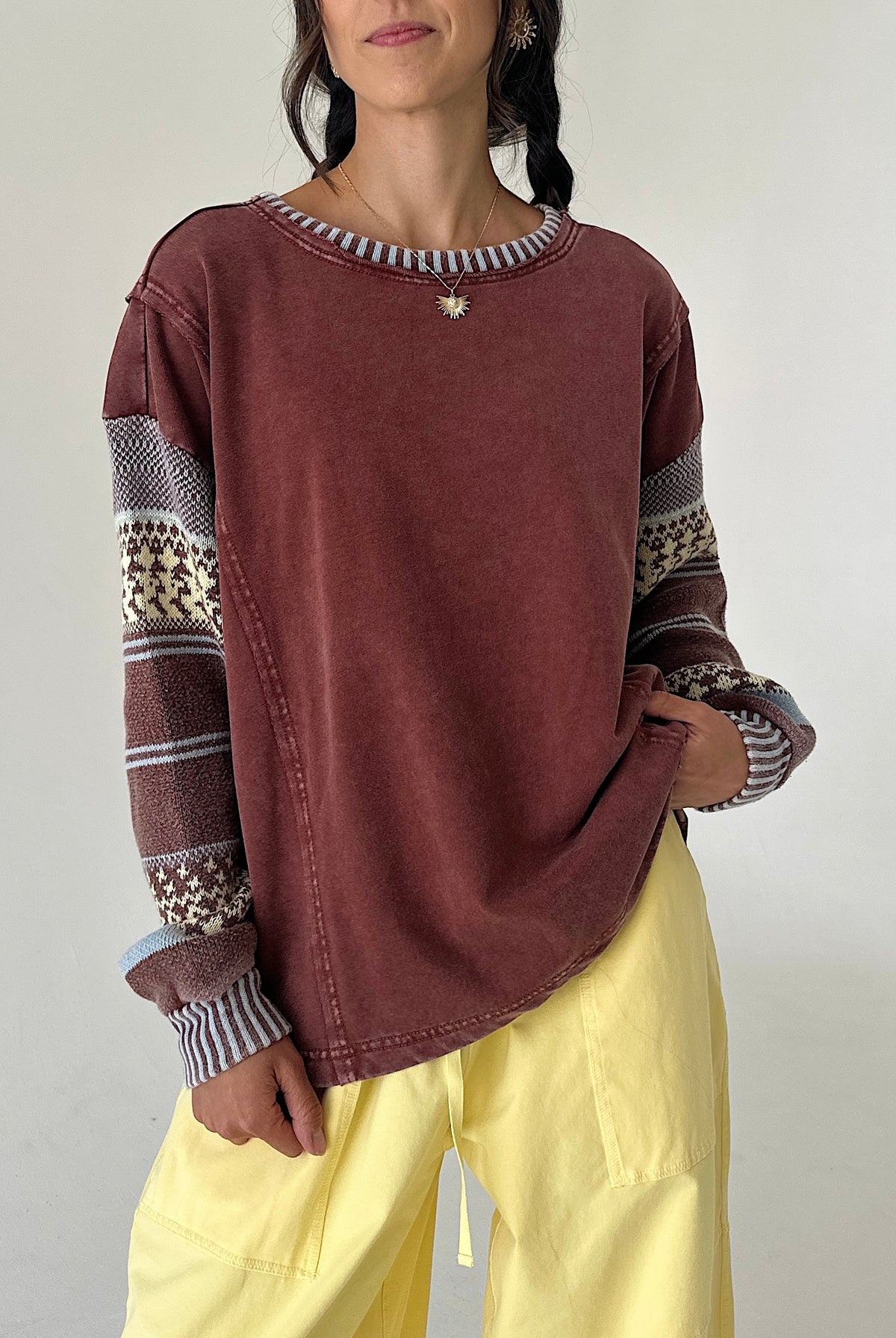 A woman wearing a washed dark reddish-brown long-sleeve sweatshirt with patterned knit cuffs and a striped crew neck, paired with light yellow pants, against a plain light gray background.