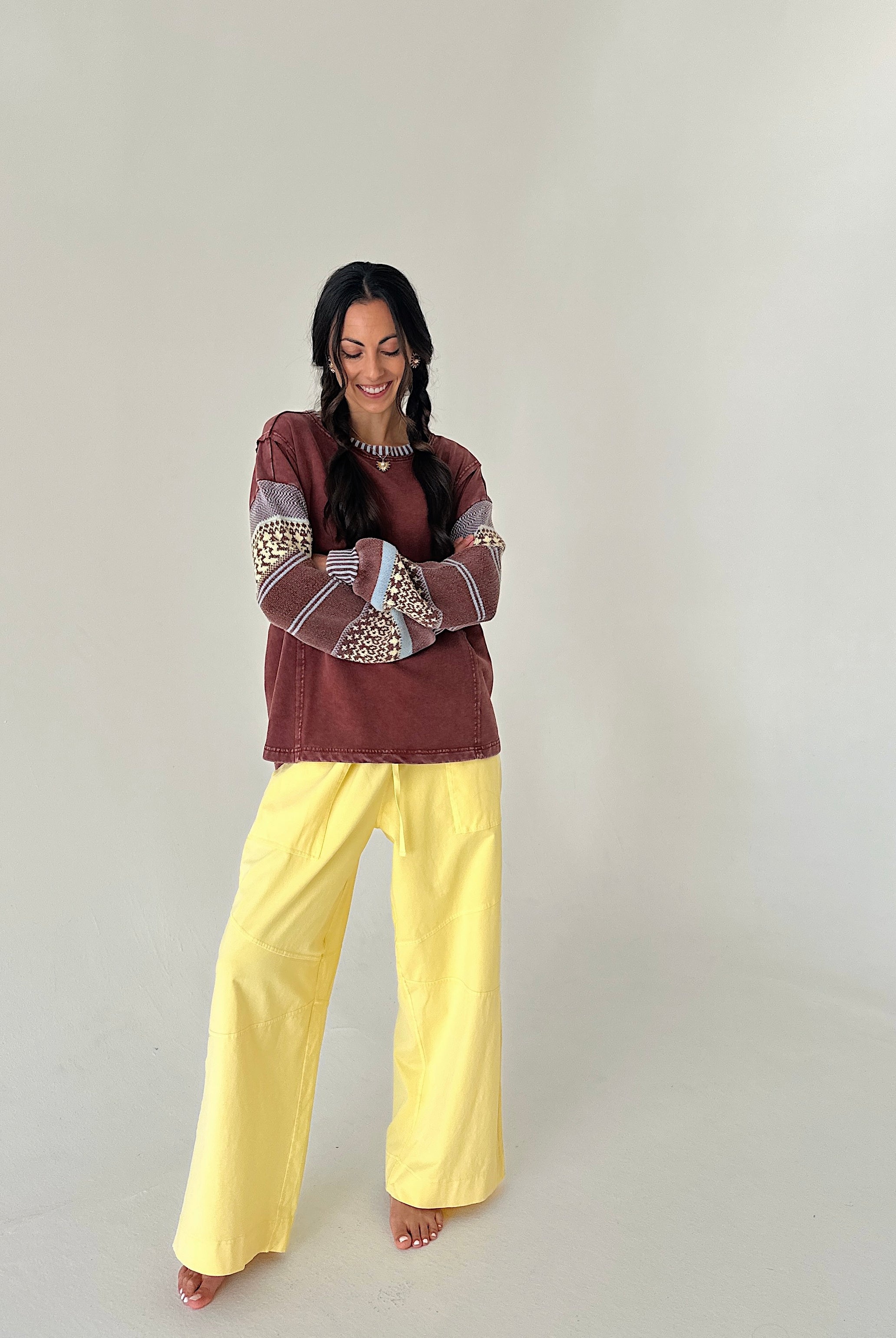 A woman modeling a bohemian-style outfit consisting of a rust-brown long-sleeve top with patterned sleeves and bright yellow wide-leg pants, standing against a plain off-white studio background.