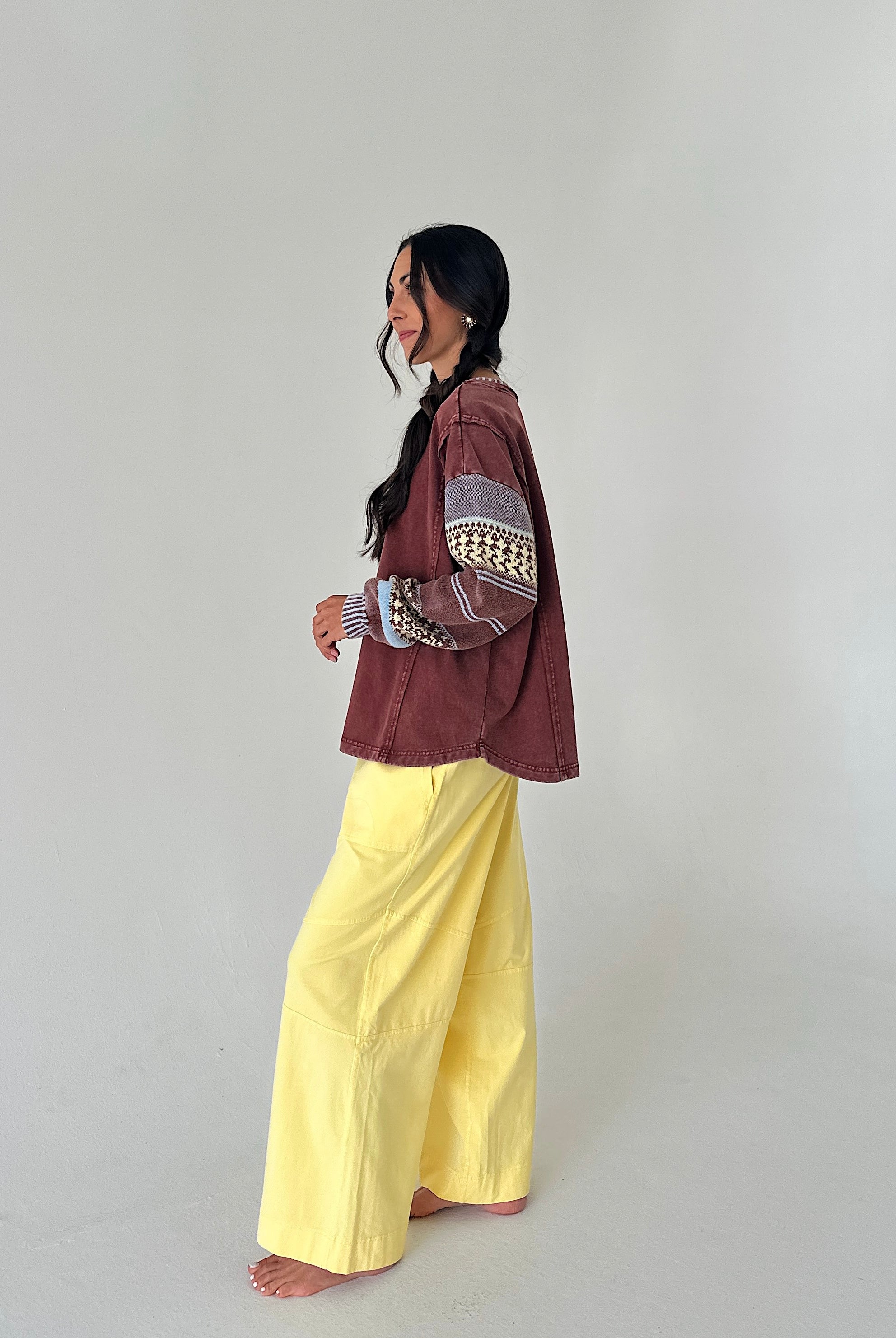 A woman stands barefoot in profile wearing a rust-brown cropped jacket with patterned knit sleeves and bright yellow wide-leg pants against a plain light gray studio background.