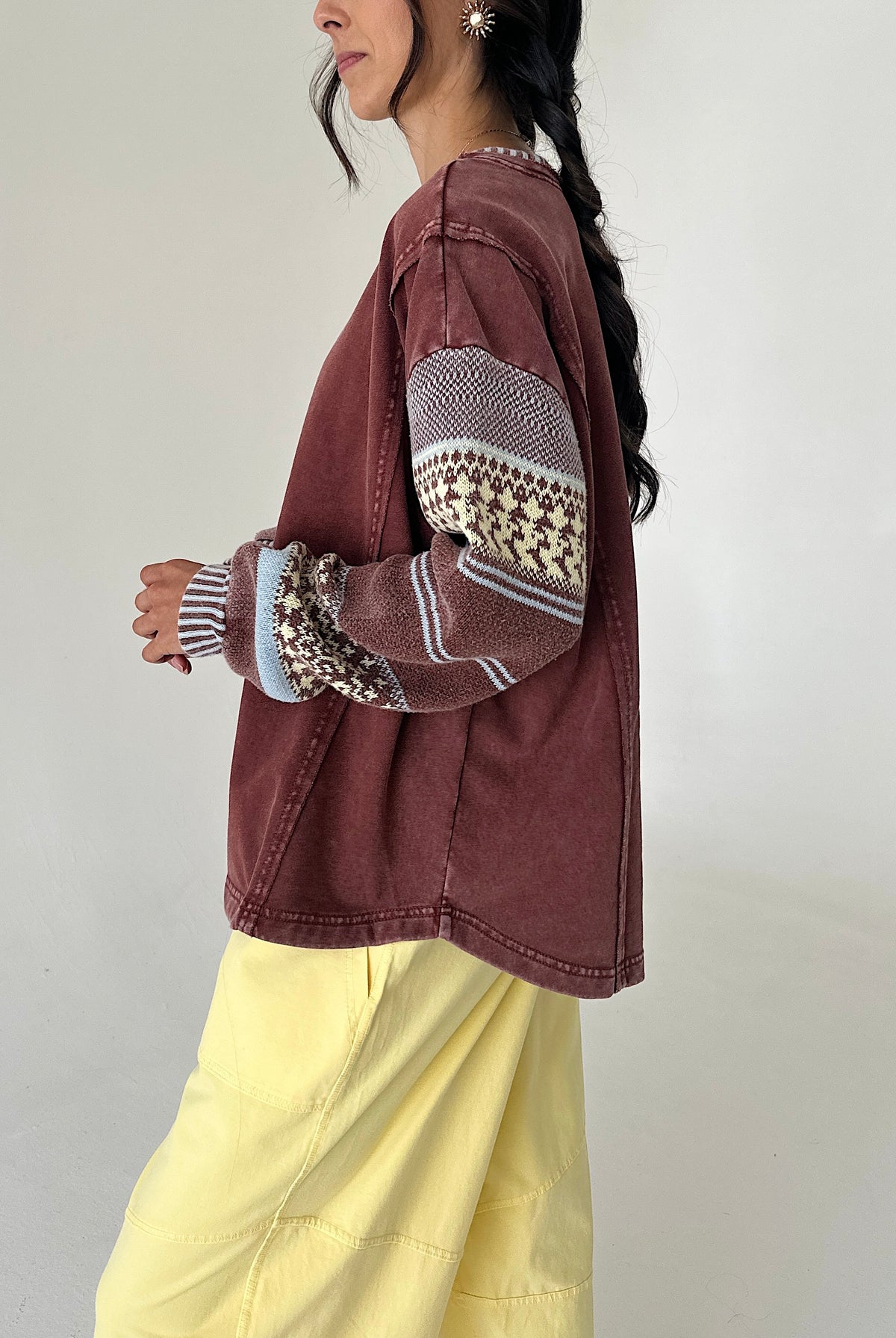 A woman wearing a washed brownish-red cropped jacket with patterned knit sleeves, paired with light yellow pants, shown from a side profile against a plain light gray wall.