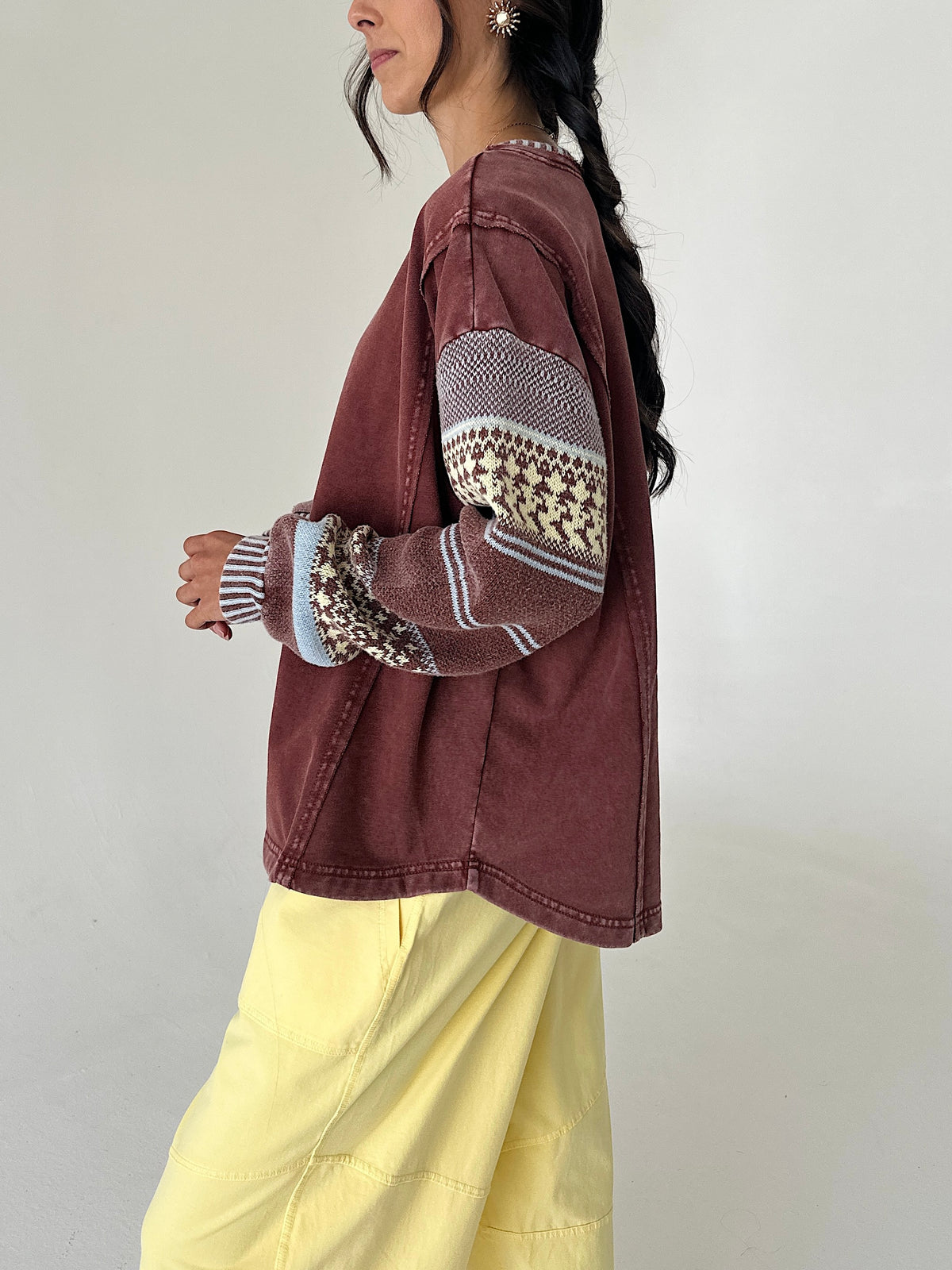A woman wearing a washed brownish-red cropped jacket with patterned knit sleeves, paired with light yellow pants, shown from a side profile against a plain light gray wall.