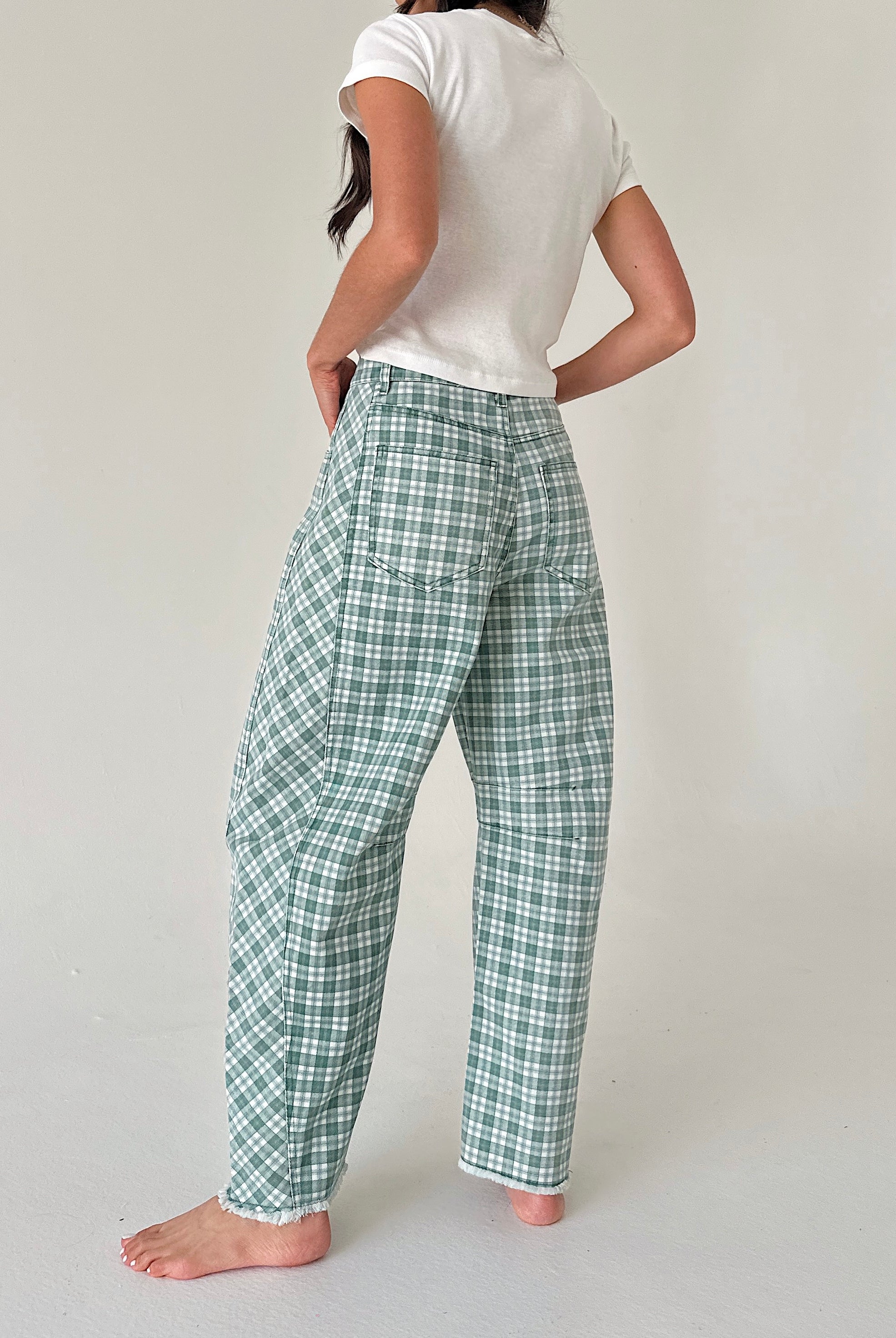 A woman shown from the back wearing green and white plaid pants, a white t-shirt, and standing barefoot against a plain light gray studio background.