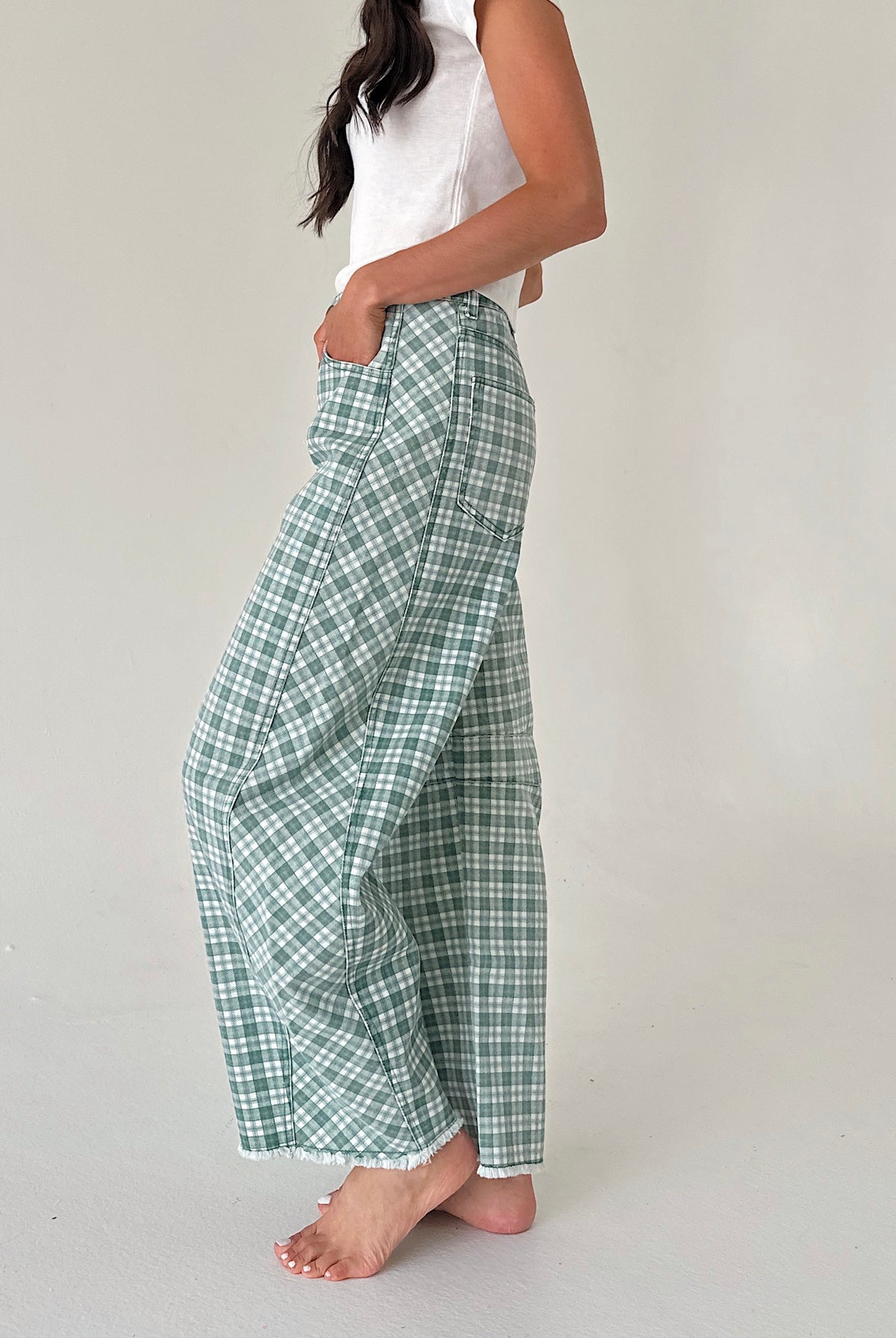 A woman wearing green and white plaid wide-leg pants with a white sleeveless top, standing barefoot against a plain light gray studio background.