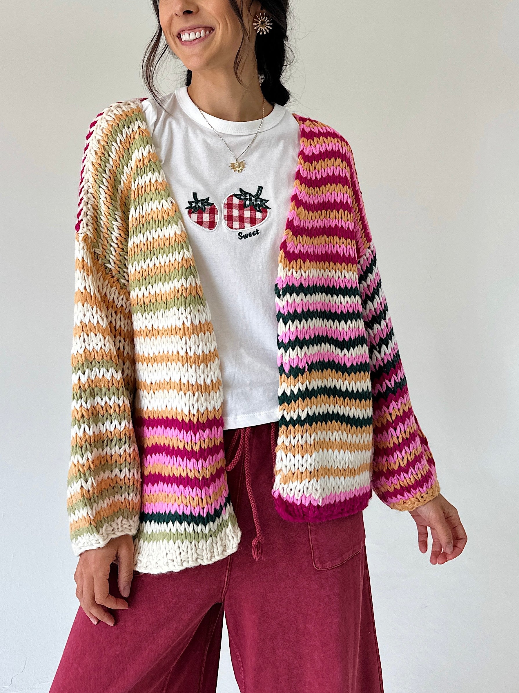A woman wearing a chunky knit cardigan with horizontal stripes in pink, orange, yellow, green, black, and white over a white graphic t-shirt and burgundy pants.