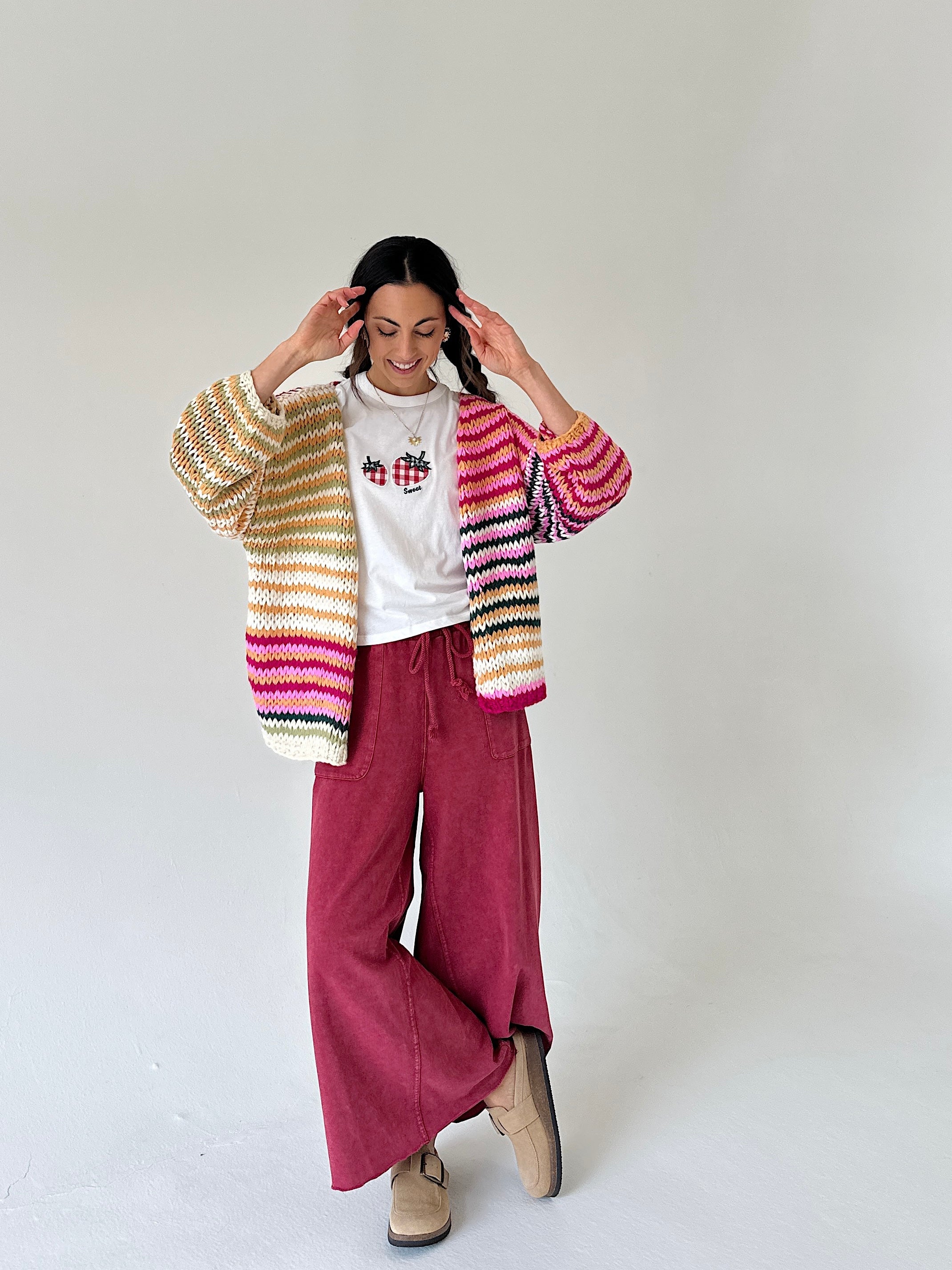 A woman modeling a colorful striped knit cardigan over a white t-shirt and magenta wide-leg pants, standing against a plain light gray studio background.