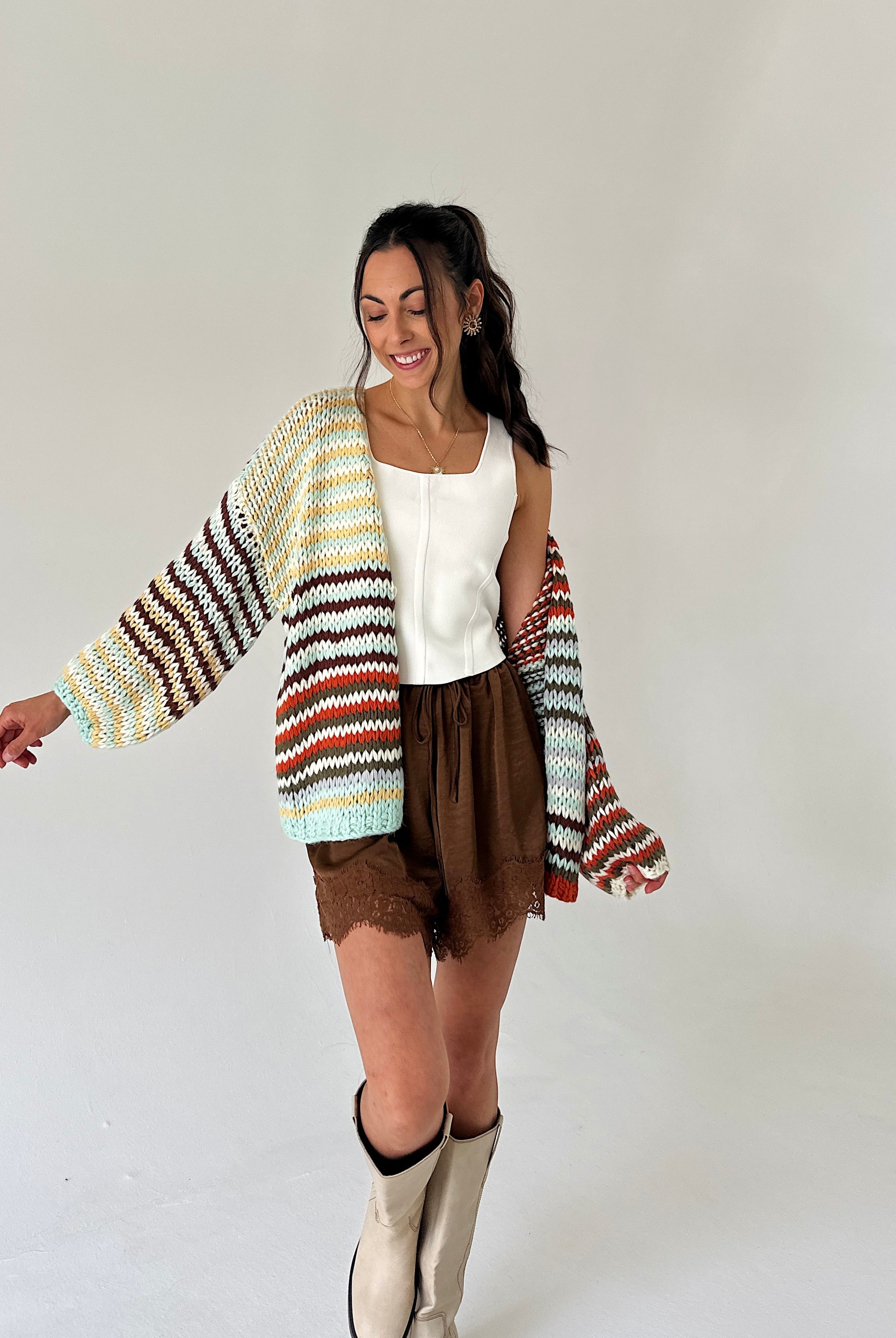 A woman modeling a chunky knit cardigan with horizontal stripes in cream, yellow, brown, orange, and light blue, worn over a white top and brown shorts.