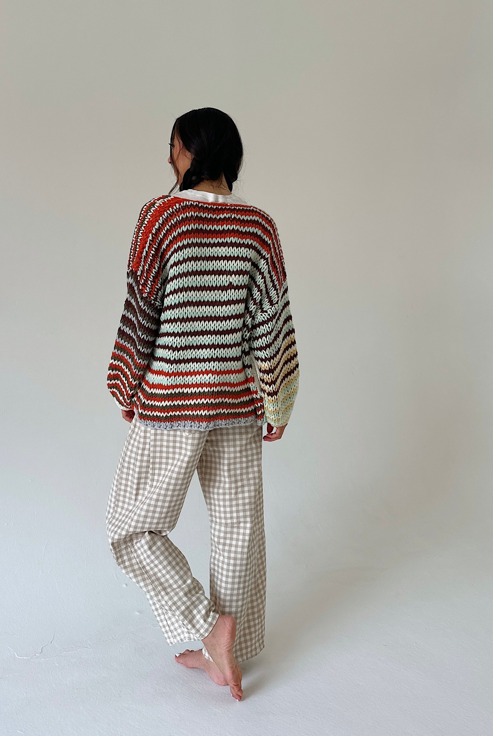 A person standing barefoot against a plain light gray studio background, viewed from the back. They are wearing a loose striped knit sweater with horizontal bands of rust red, cream, brown, and black, paired with light beige gingham pants.