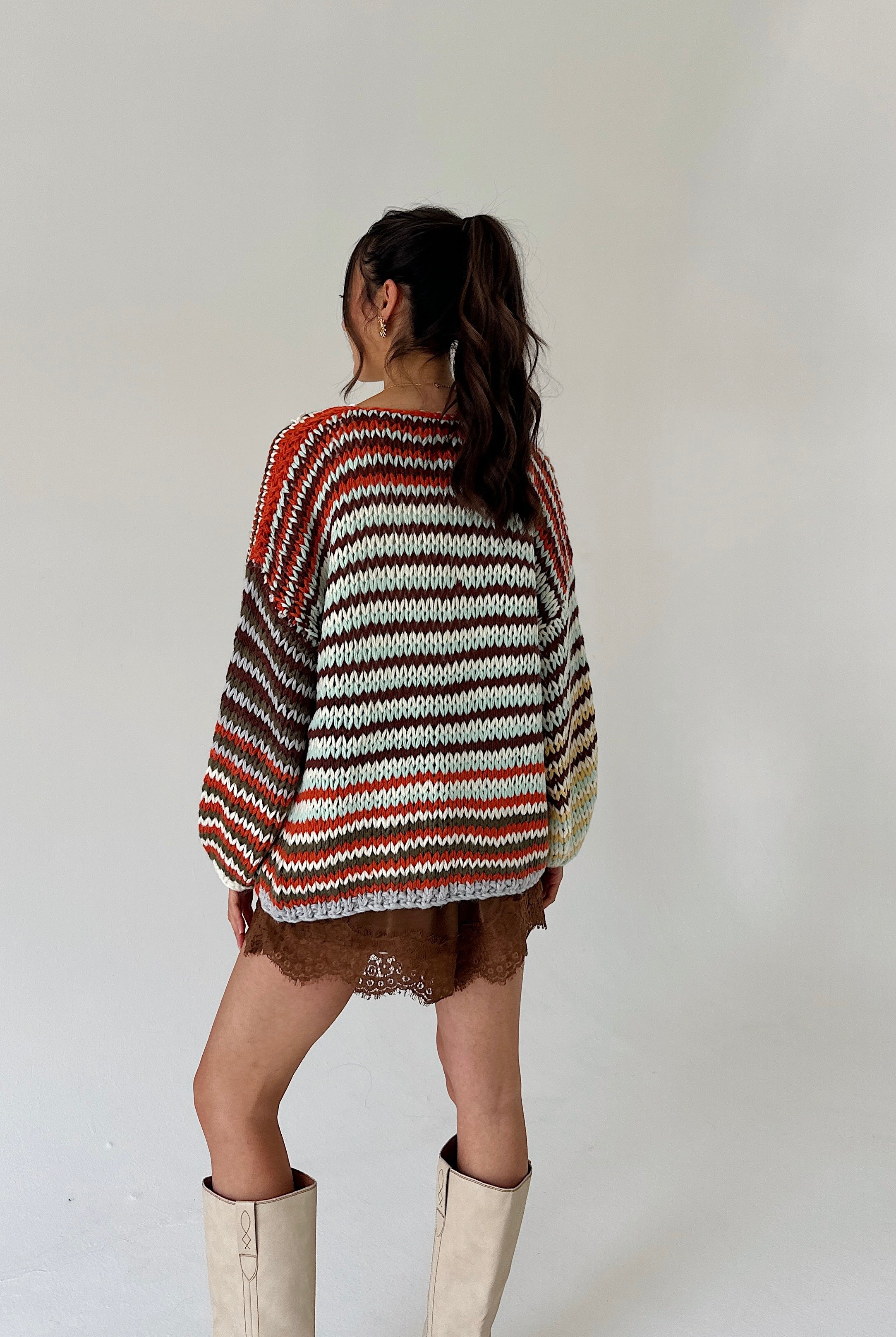 A woman is shown from behind wearing a chunky knit sweater with horizontal stripes in rust orange, brown, cream, and dark brown, paired with brown shorts and off-white boots, against a plain light gray background.