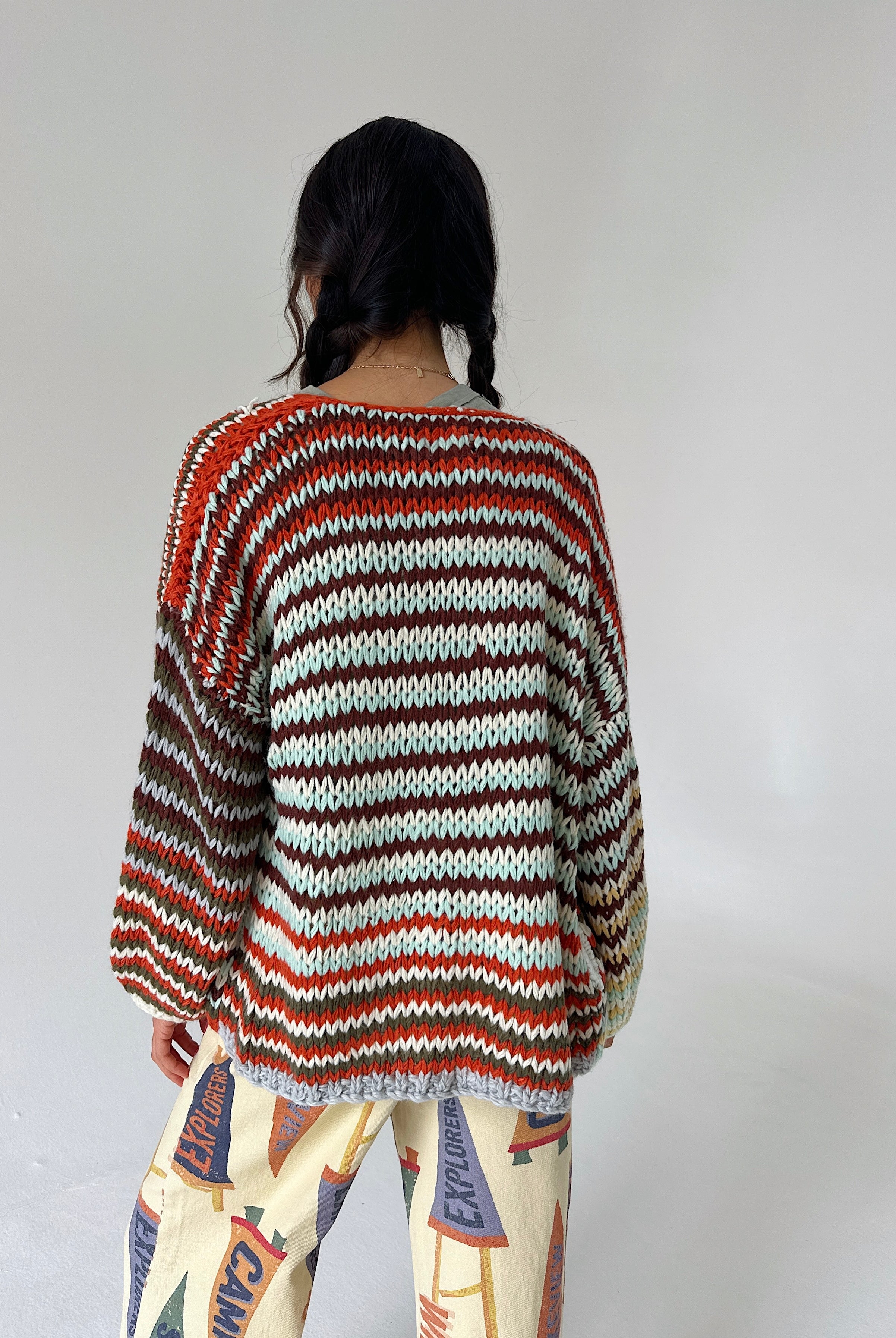 A person shown from the back wearing a loose, long-sleeve striped knit sweater with horizontal bands of rust red, dark brown, cream, and gray, paired with patterned pants against a plain light gray studio background.