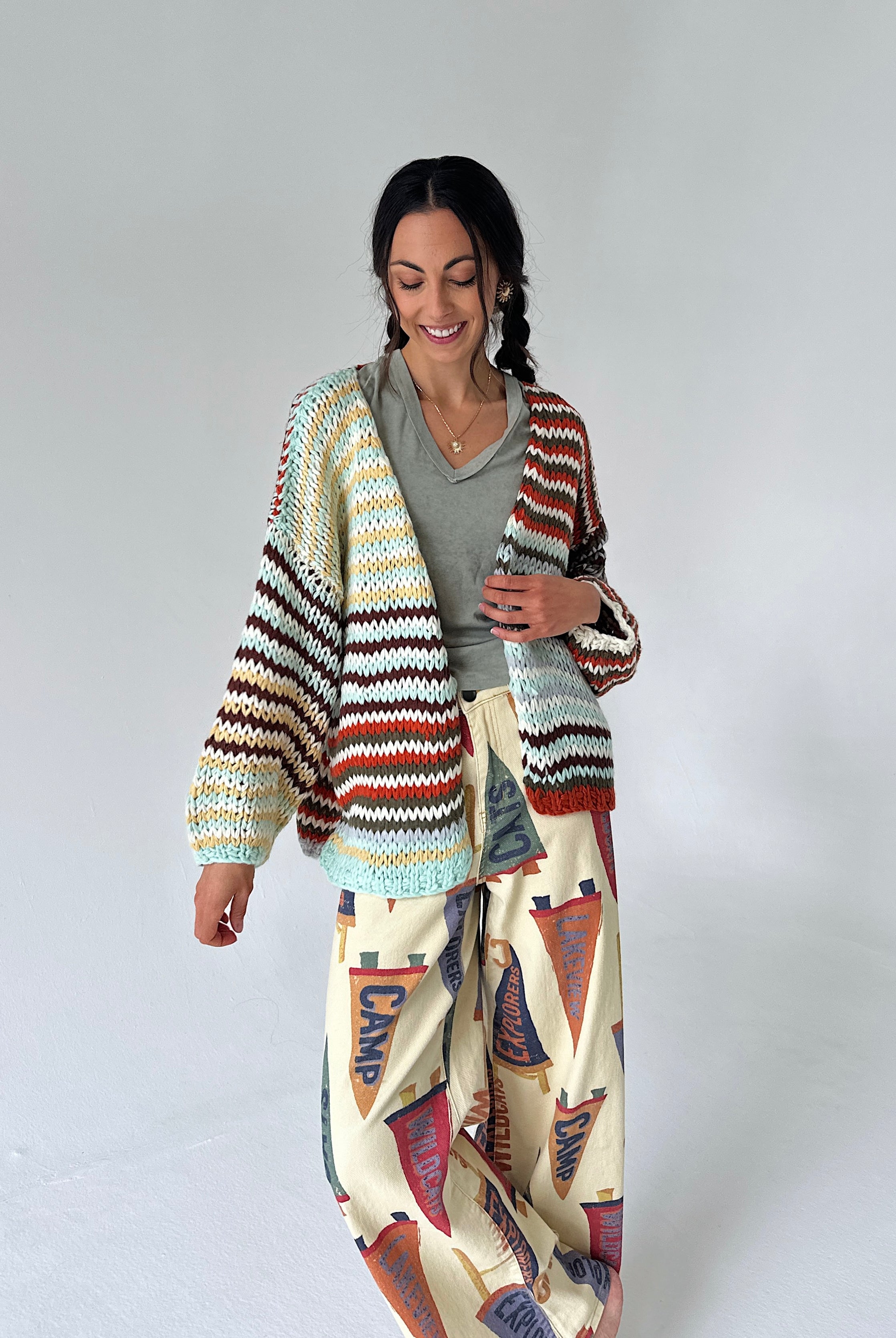 A woman modeling a chunky knit cardigan with horizontal multicolor stripes in beige, brown, red, and light blue, worn over a gray top and paired with cream wide-leg pants featuring a red, blue, and brown flag pattern.