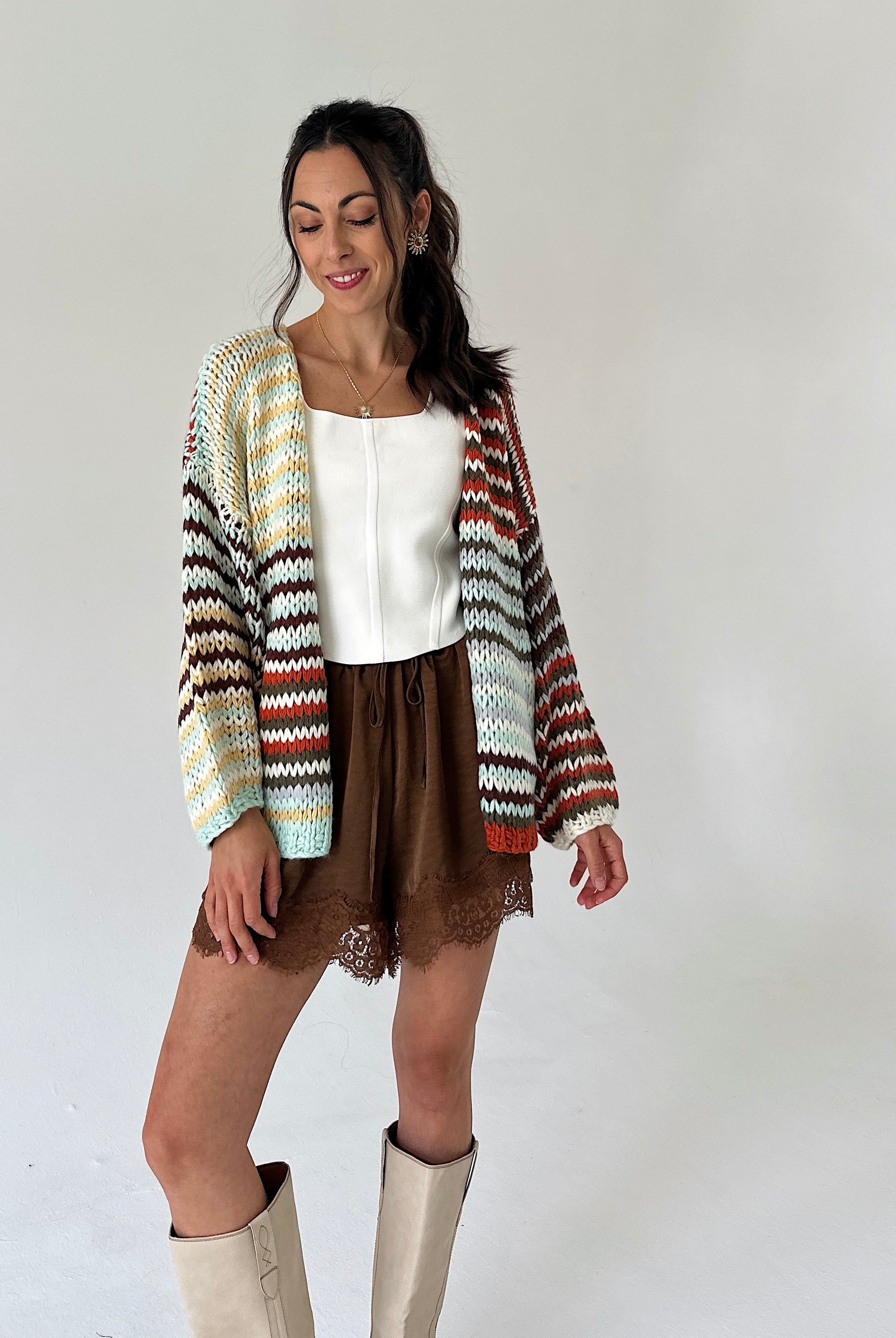 A woman modeling a long-sleeve knit cardigan with a multicolor chevron pattern in cream, orange, brown, and green, worn over a white top and brown shorts.