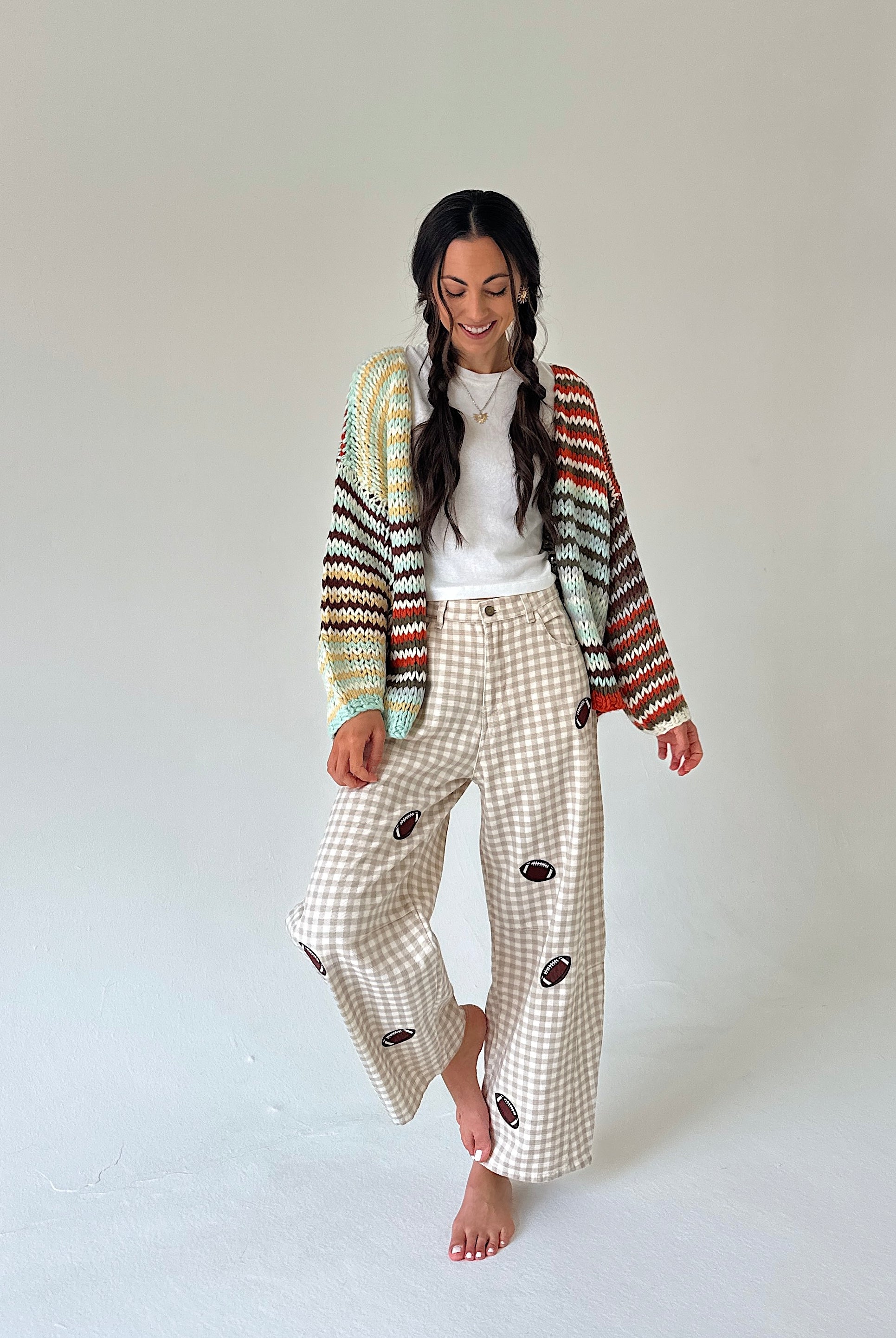 A woman stands barefoot against a plain light gray studio background, wearing a cream gingham wide-leg pant set with dark brown football patches, a white top, and a multicolor striped knit cardigan.