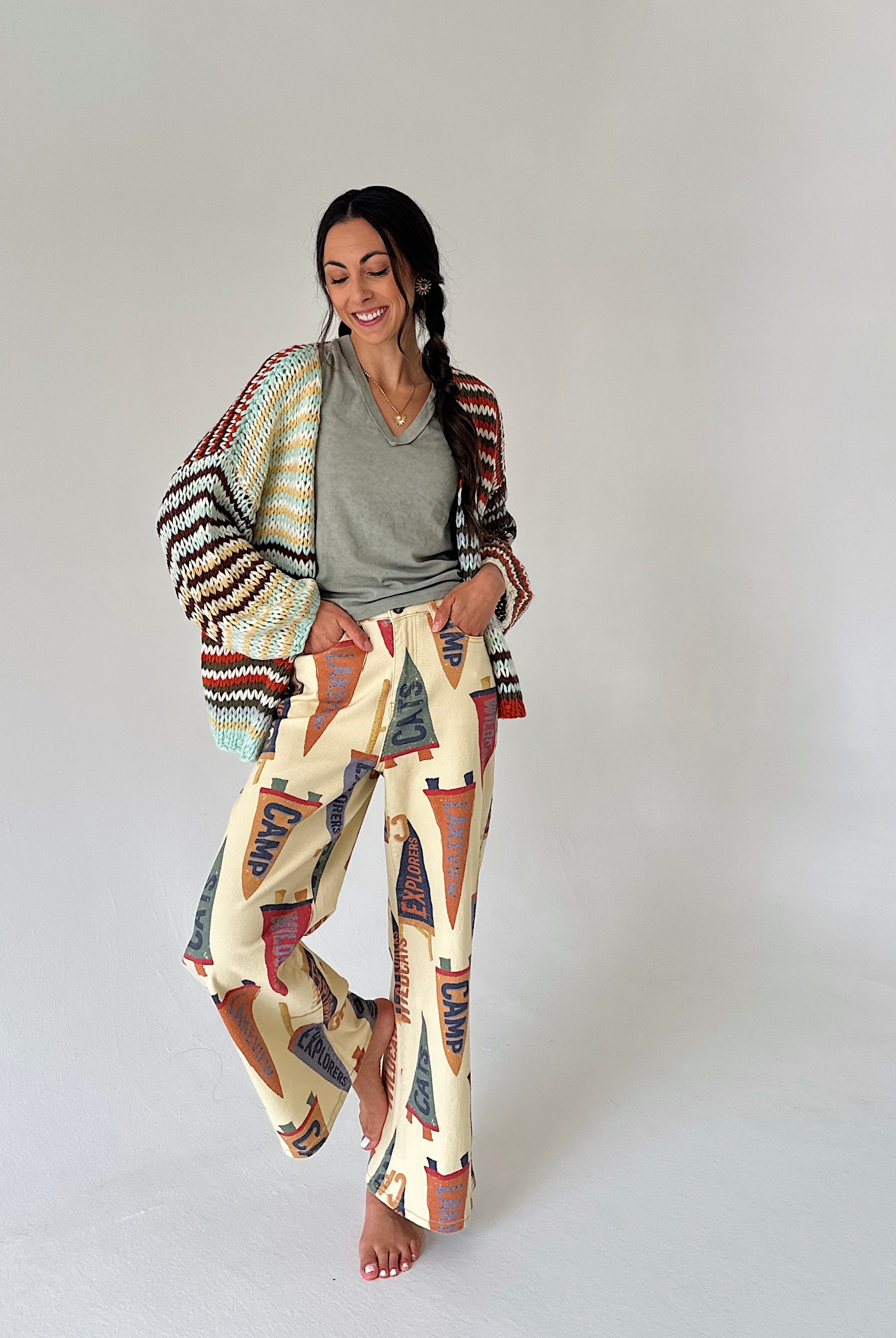 A woman modeling a bohemian-style outfit consisting of wide-leg cream pants with a colorful penant print, a gray V-neck t-shirt, and a striped knit cardigan draped over her shoulders, set against a plain light gray studio background.