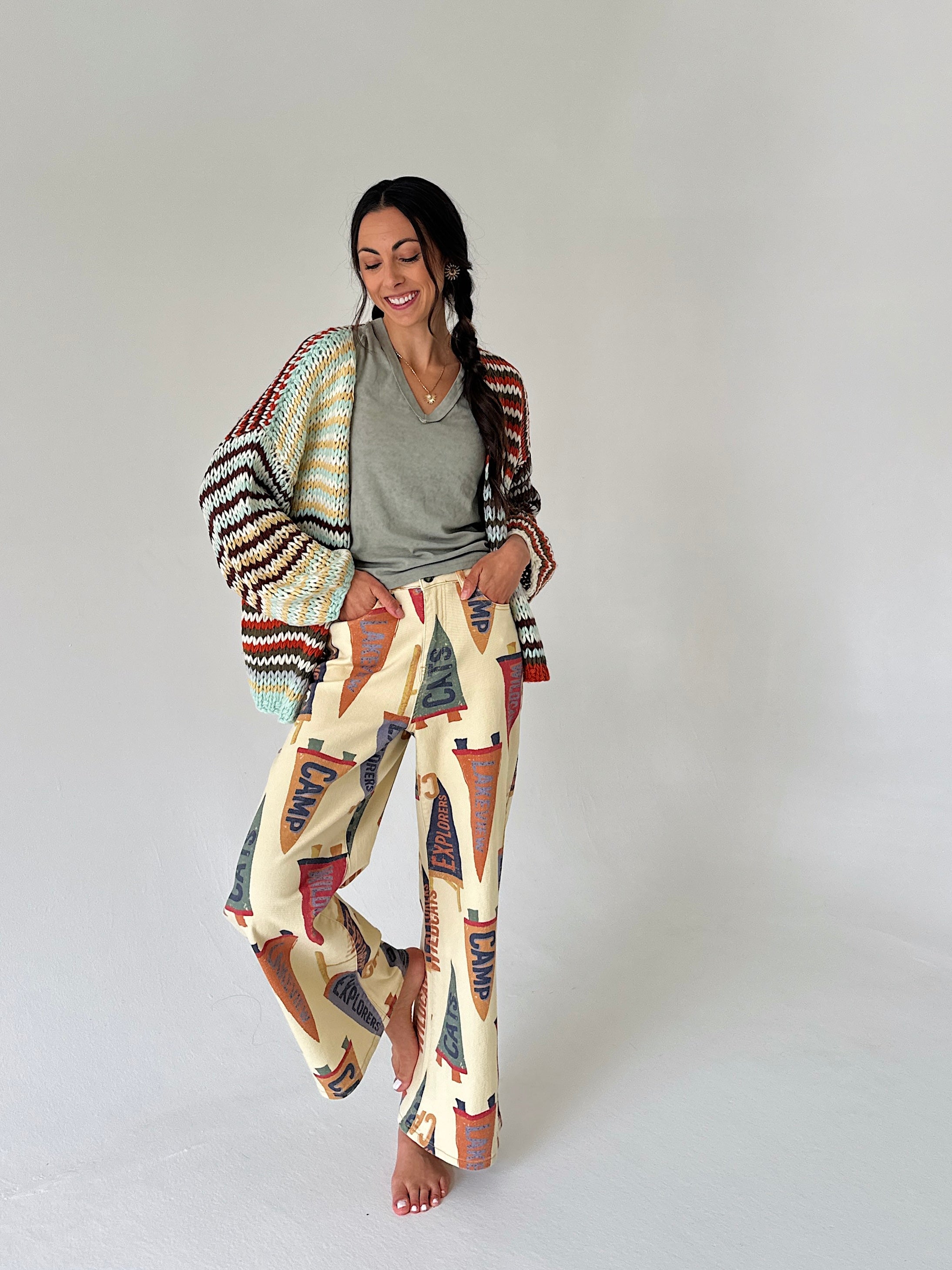 A woman modeling a bohemian-style outfit consisting of wide-leg cream pants with a colorful penant print, a gray V-neck t-shirt, and a striped knit cardigan draped over her shoulders, set against a plain light gray studio background.