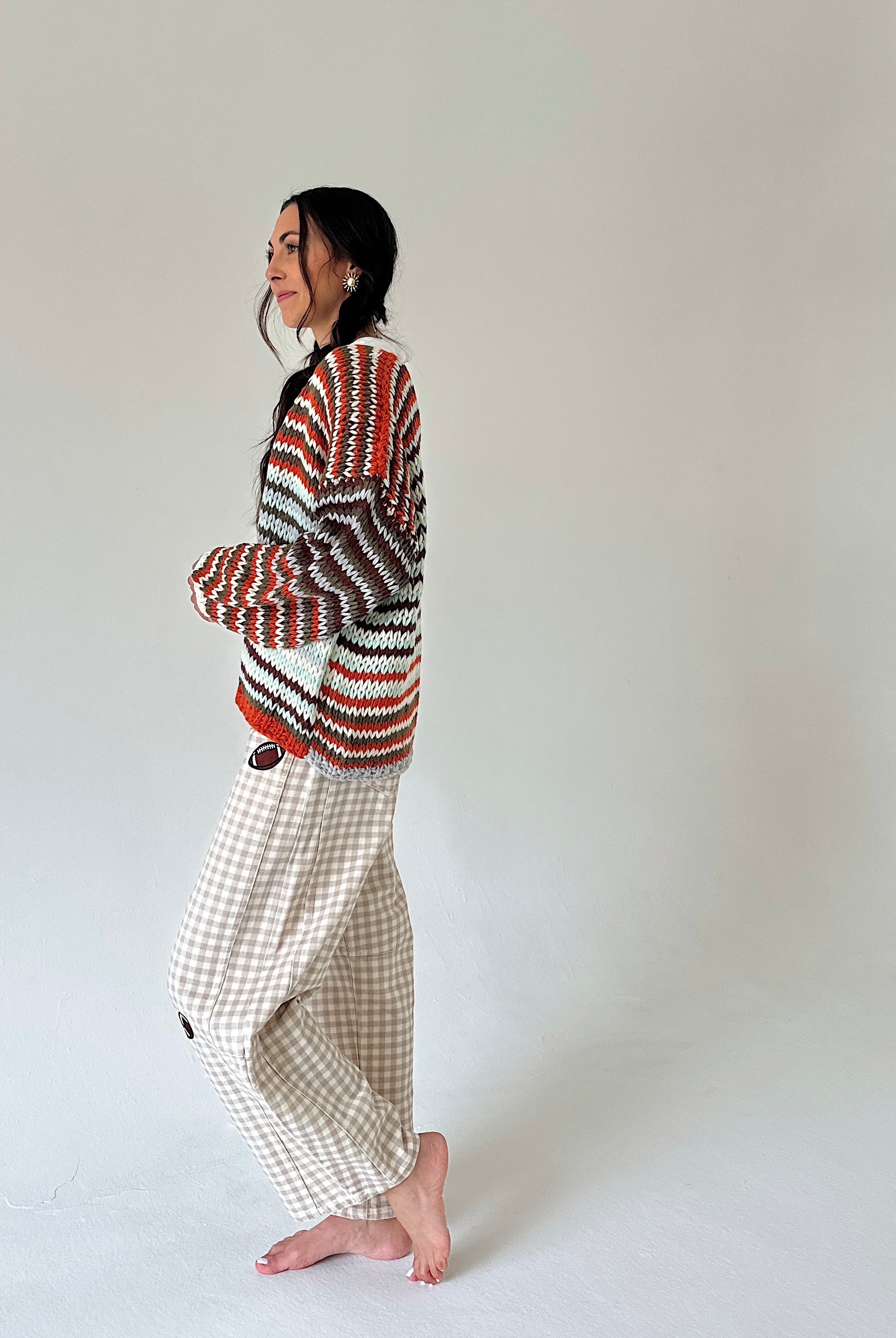 A woman stands barefoot in profile against a plain light gray studio background, wearing a multicolor knit sweater with red, orange, black, and white stripes and beige gingham pants.