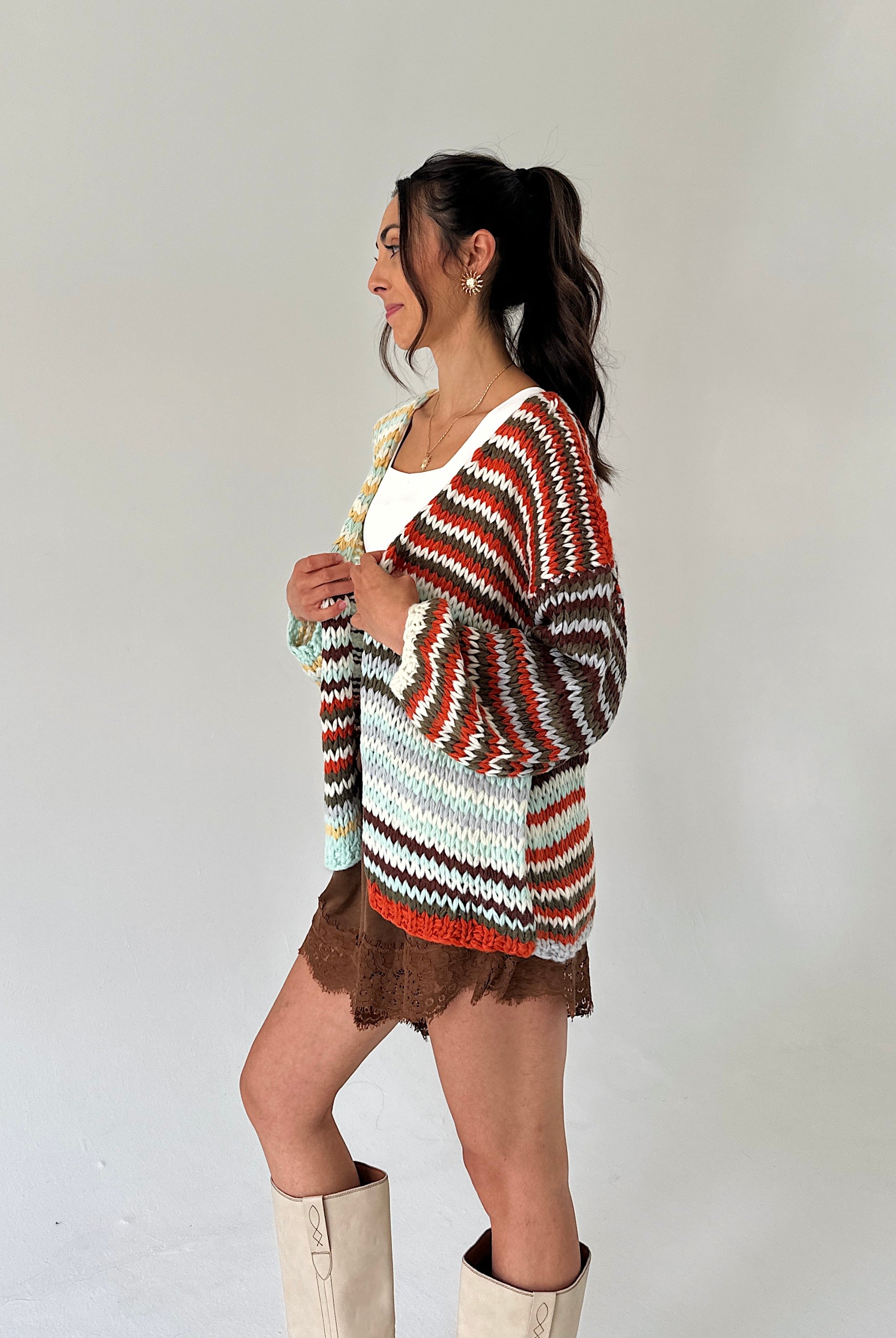 A woman modeling a chunky knit cardigan with horizontal wavy stripes in rust orange, brown, white, and mint green, styled with a white top, brown skirt, and beige boots against a plain light gray background.