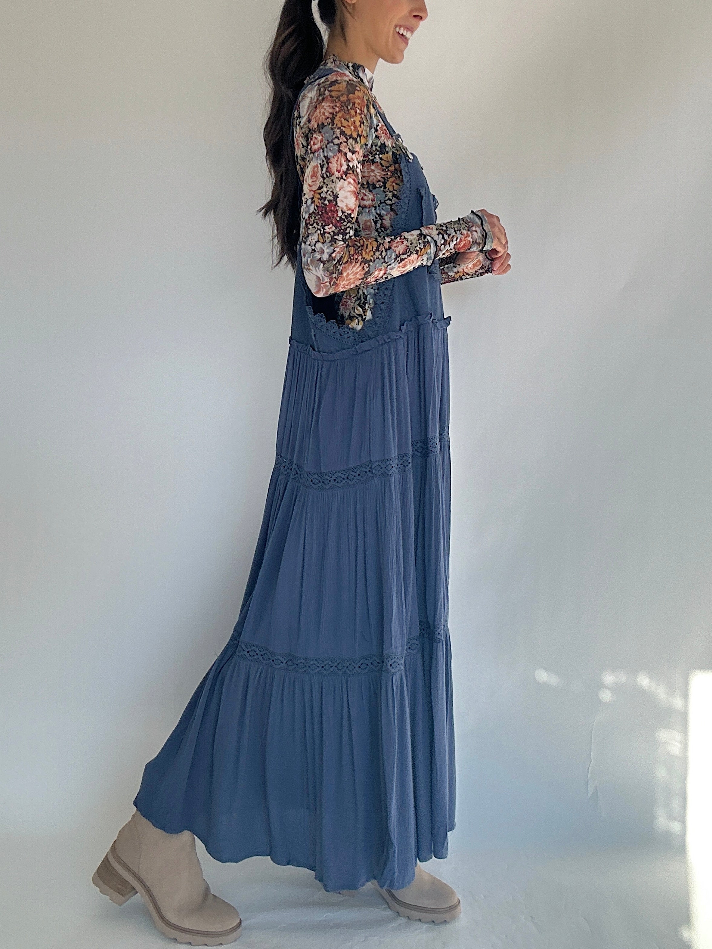 Woman wearing a blue overall dress with floral long sleeve top against a white background showing side view