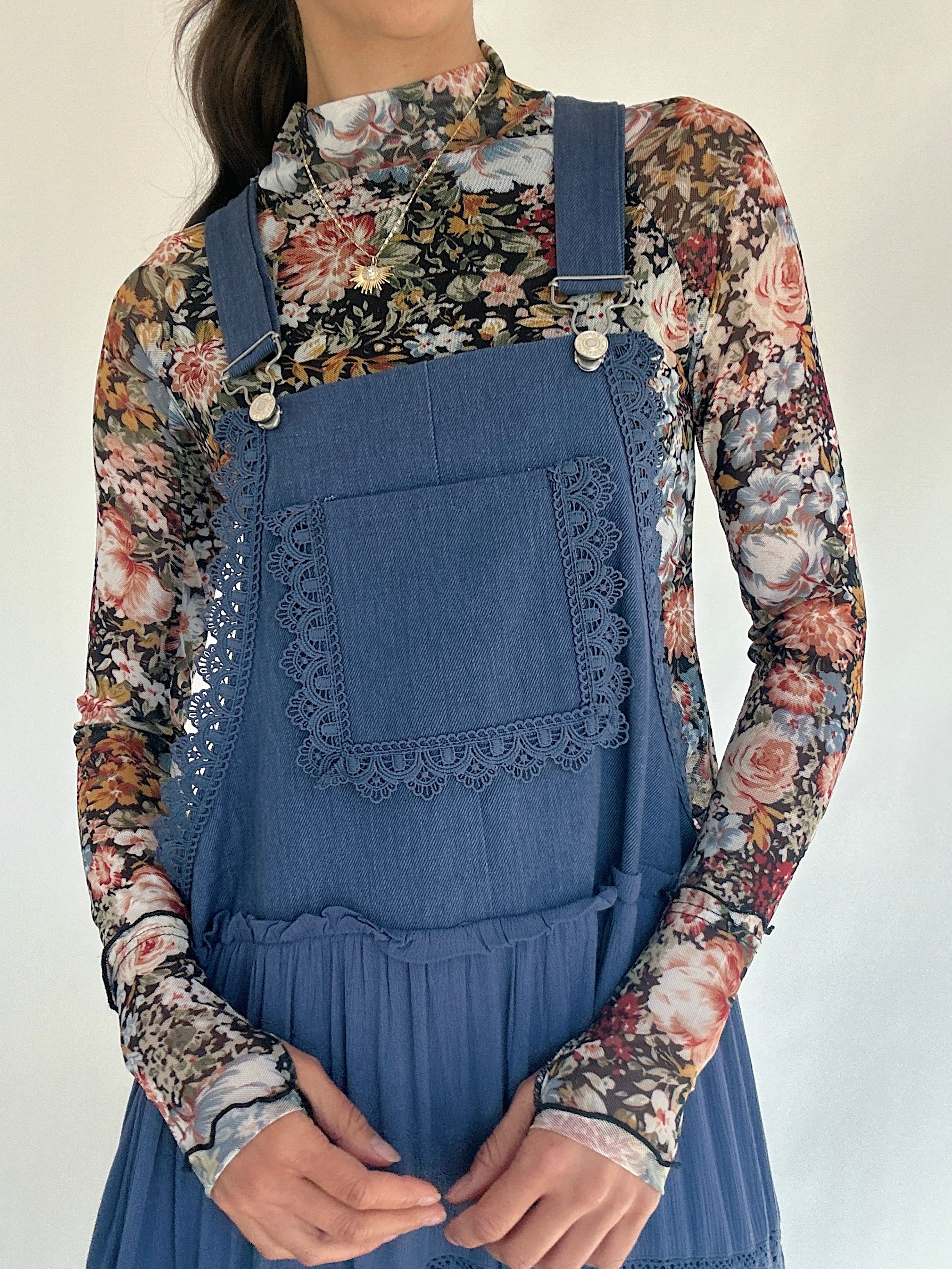 Person wearing a floral shirt under a blue denim overall dress with lace detailing on a plain background